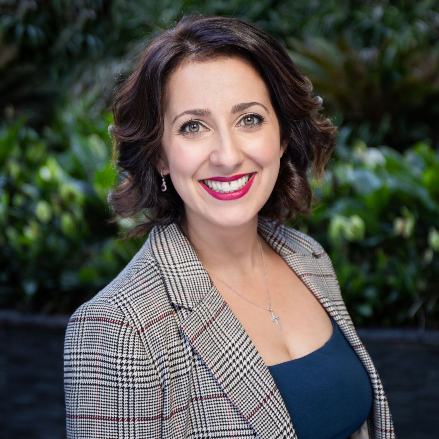 Professional headshot against natural outdoor background wearing plaid blazer.