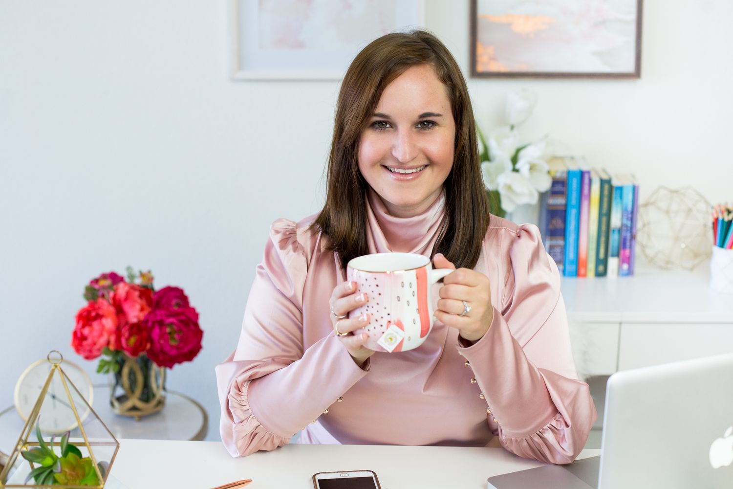 Professional wearing a pink sweater holds a coffee mug while working at a bright desk with flowers.