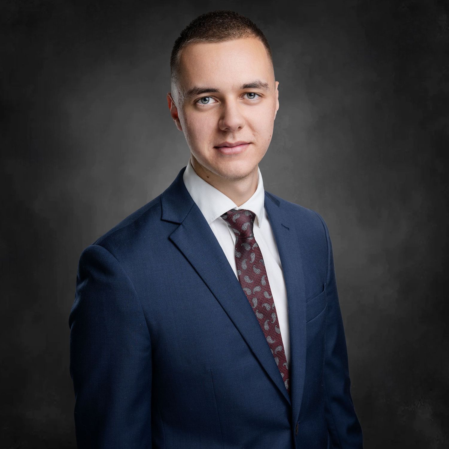 Young professional in navy suit and patterned tie photographed against dark background.