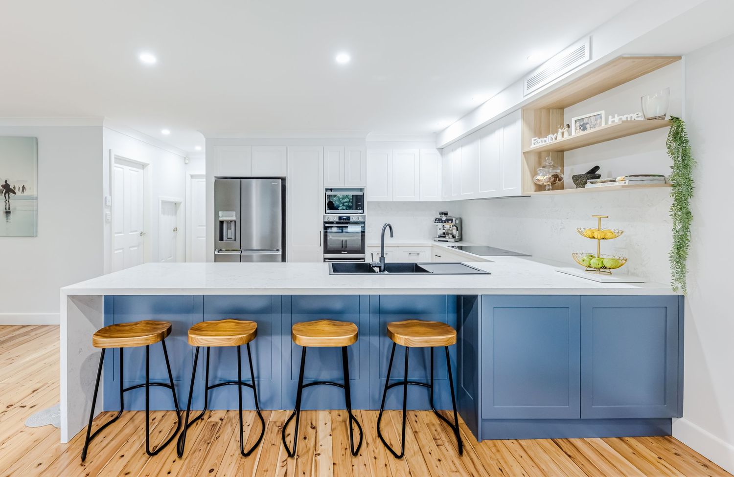 Modern kitchen featuring blue base cabinets, white countertops, wooden bar stools, and open shelving with recessed lighting.