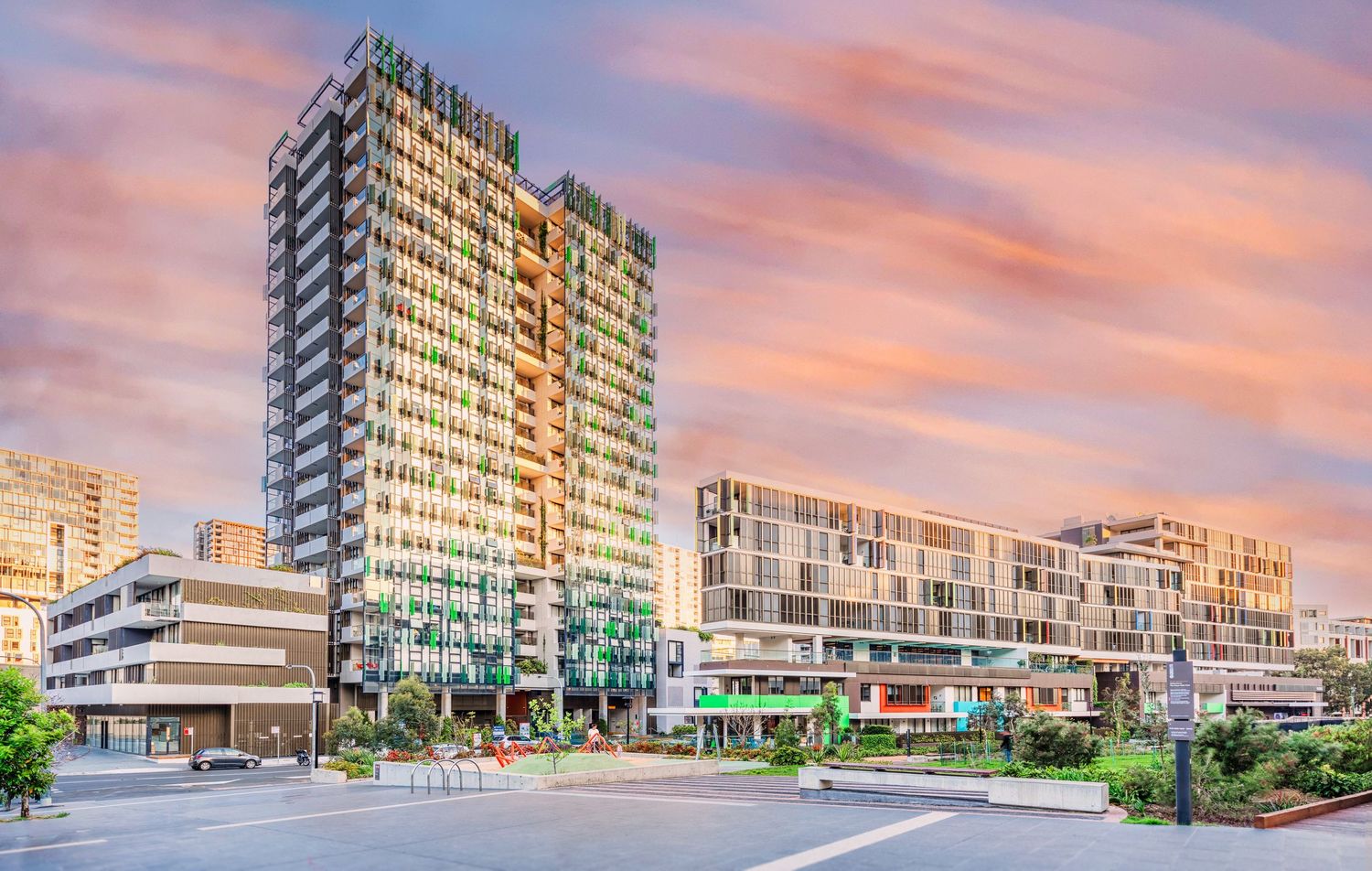 Modern apartment buildings with glass facades reflect a vibrant pink and orange sunset sky along an urban waterfront.
