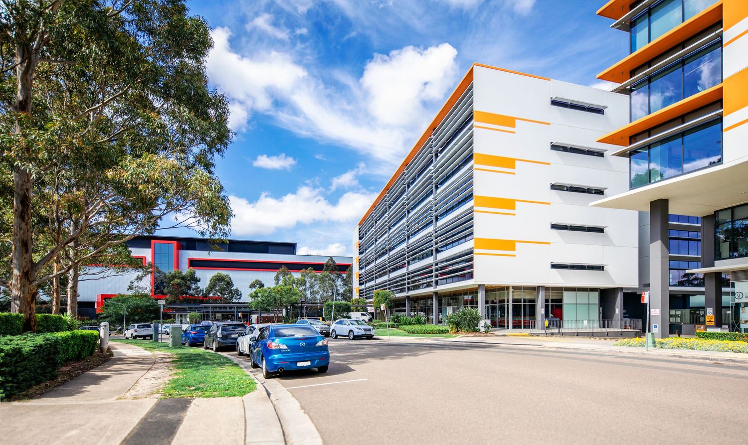 Modern office building exterior with orange accents and glass windows along a tree-lined walkway on a sunny day.