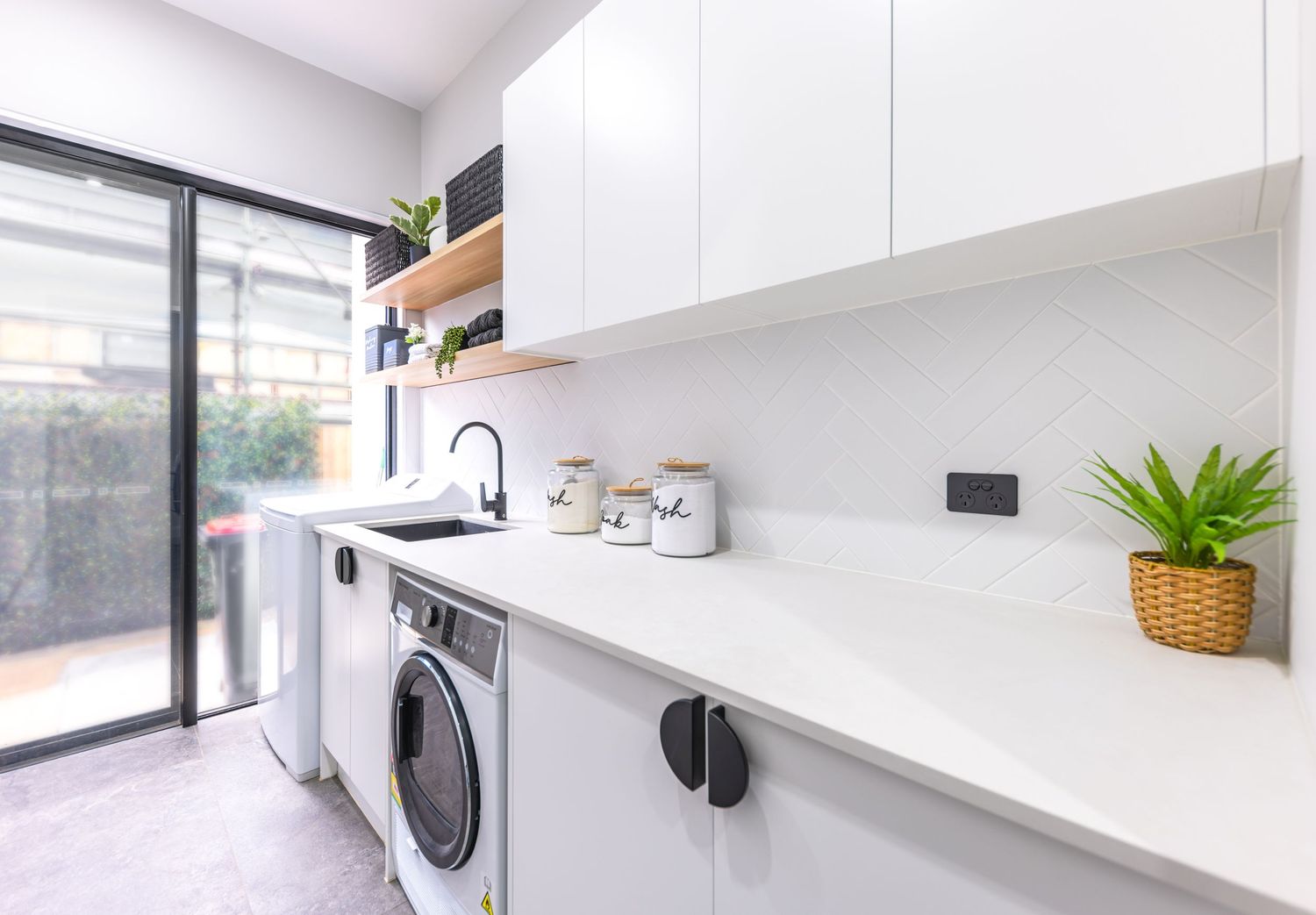 Modern white laundry room with built-in cabinets, washing machine, and potted plant near glass sliding door.