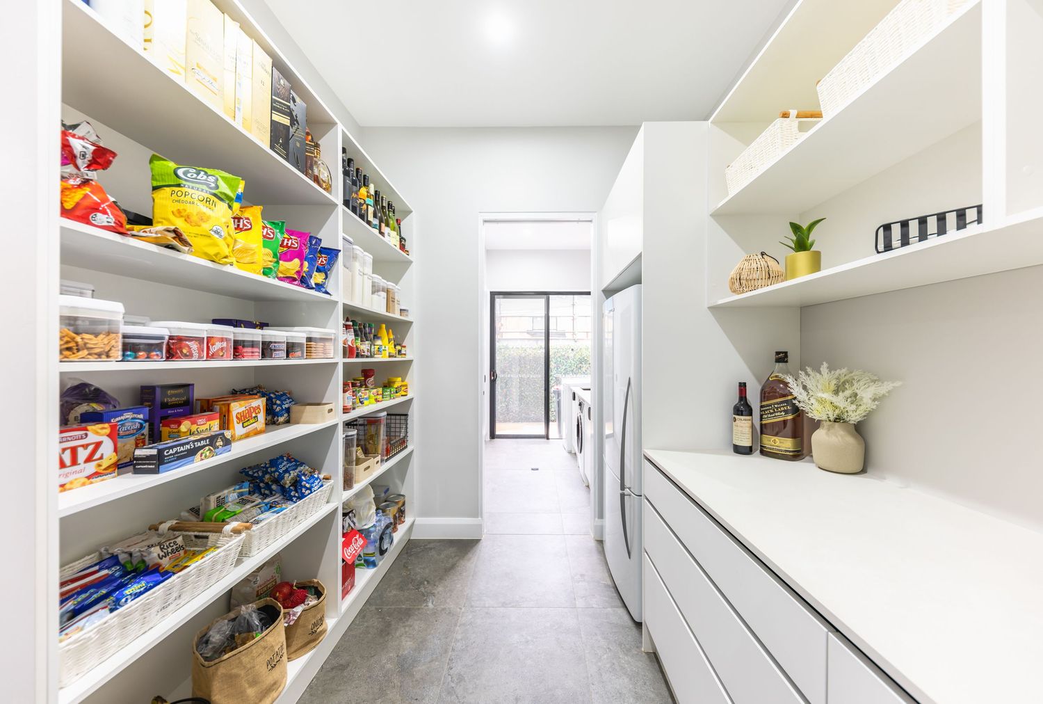 Long white pantry with built-in shelving units displaying organized food items and household supplies along both walls.