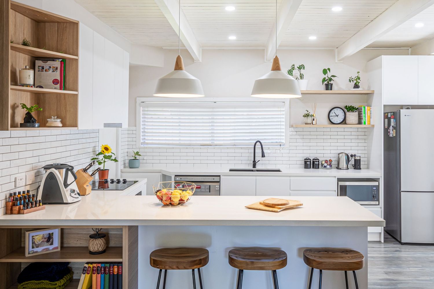 Modern white kitchen with wood bar stools, pendant lights, and open shelving features a large center island.