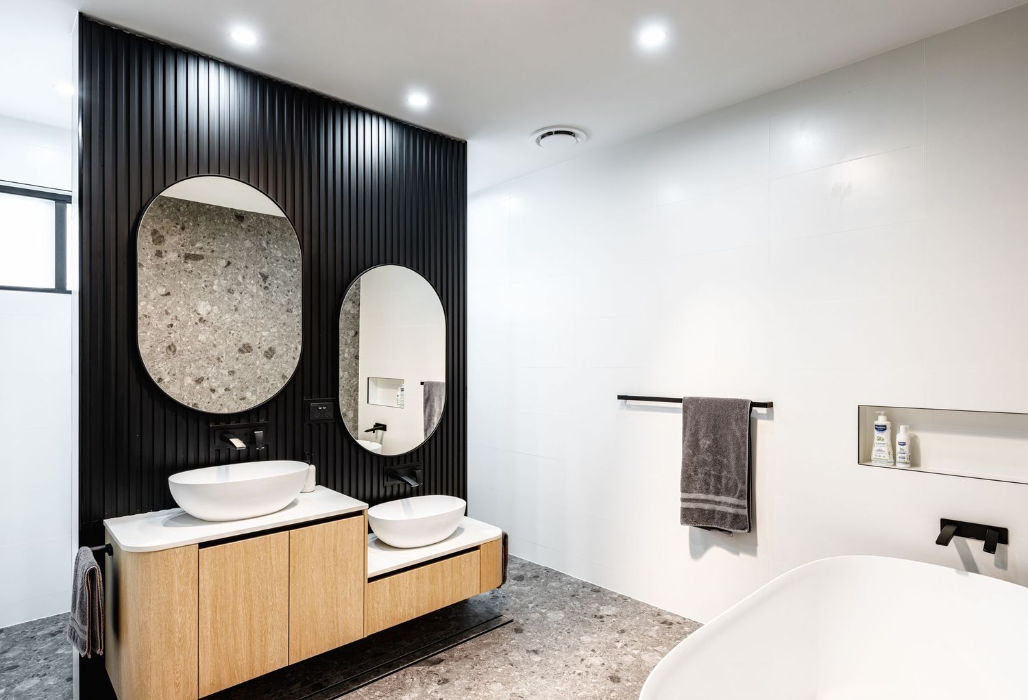 Modern bathroom interior with black accent wall, round mirrors, wooden vanity, and recessed lighting.