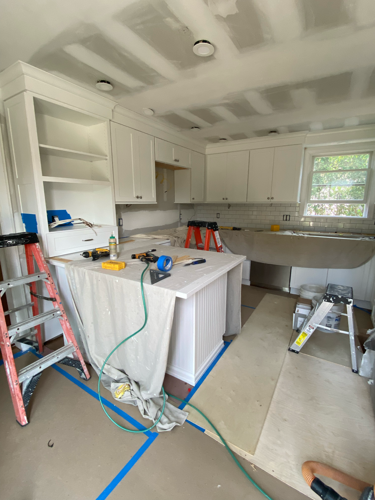 Kitchen renovation in progress with white cabinets, ladders, tools and blue painter's tape on floor showing work underway.