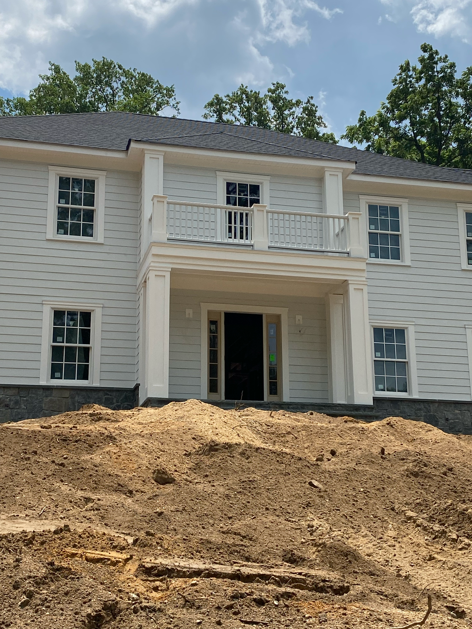 A white colonial house with black shutters sits behind a large mound of excavated dirt during construction work.