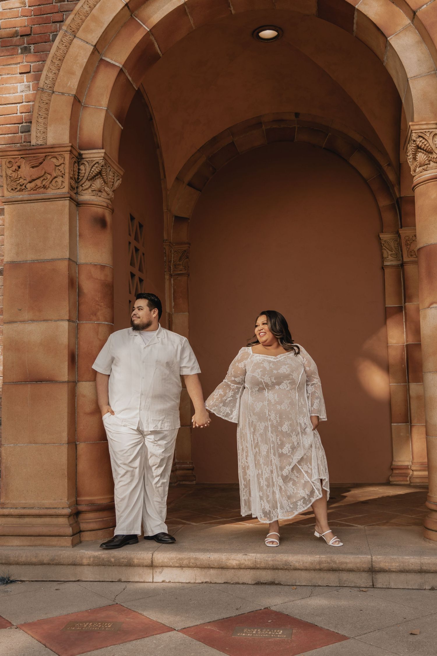 A couple poses under brick archways in matching white outfits.