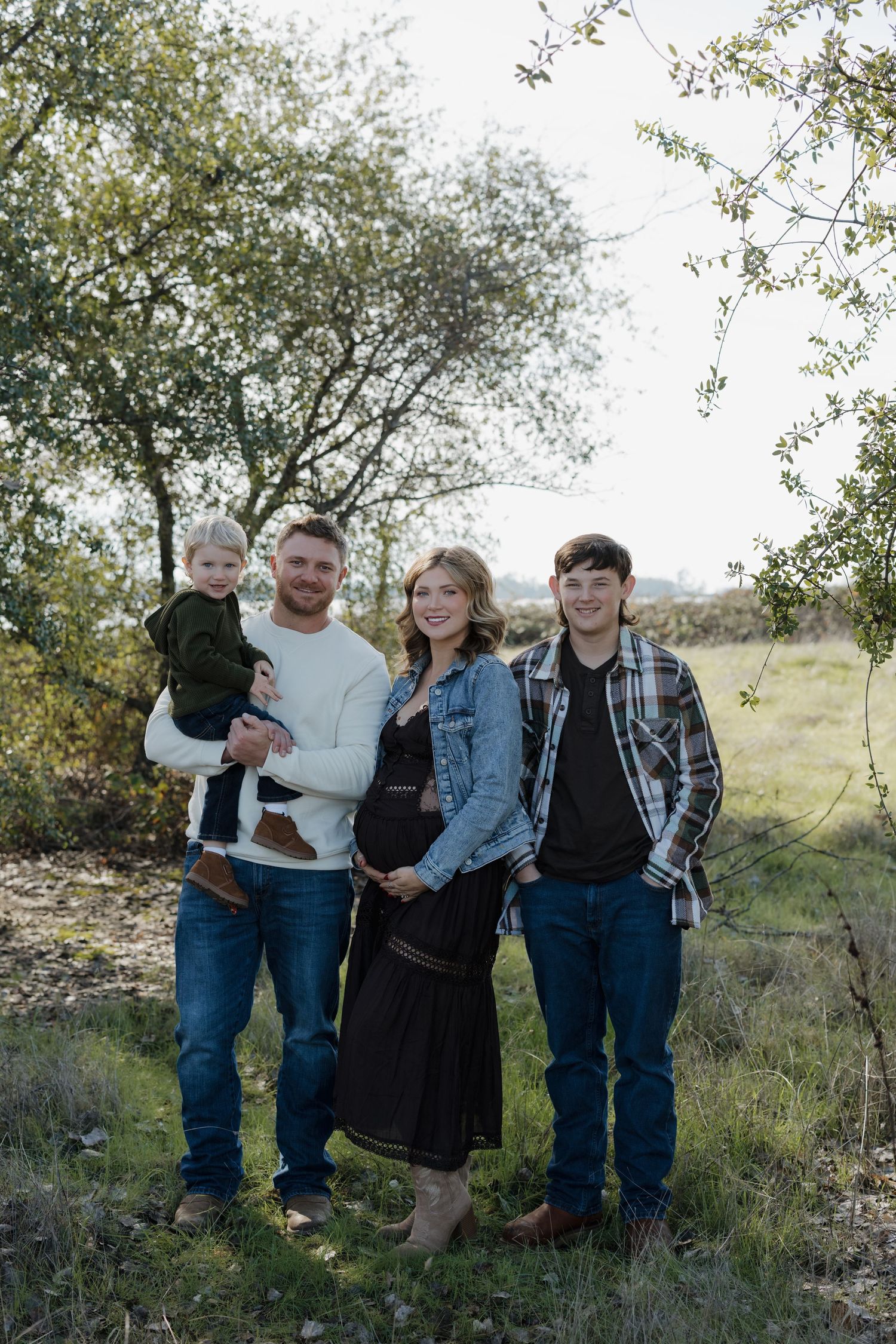 A family stands together outdoors in casual clothing against a backdrop of trees and natural scenery.
