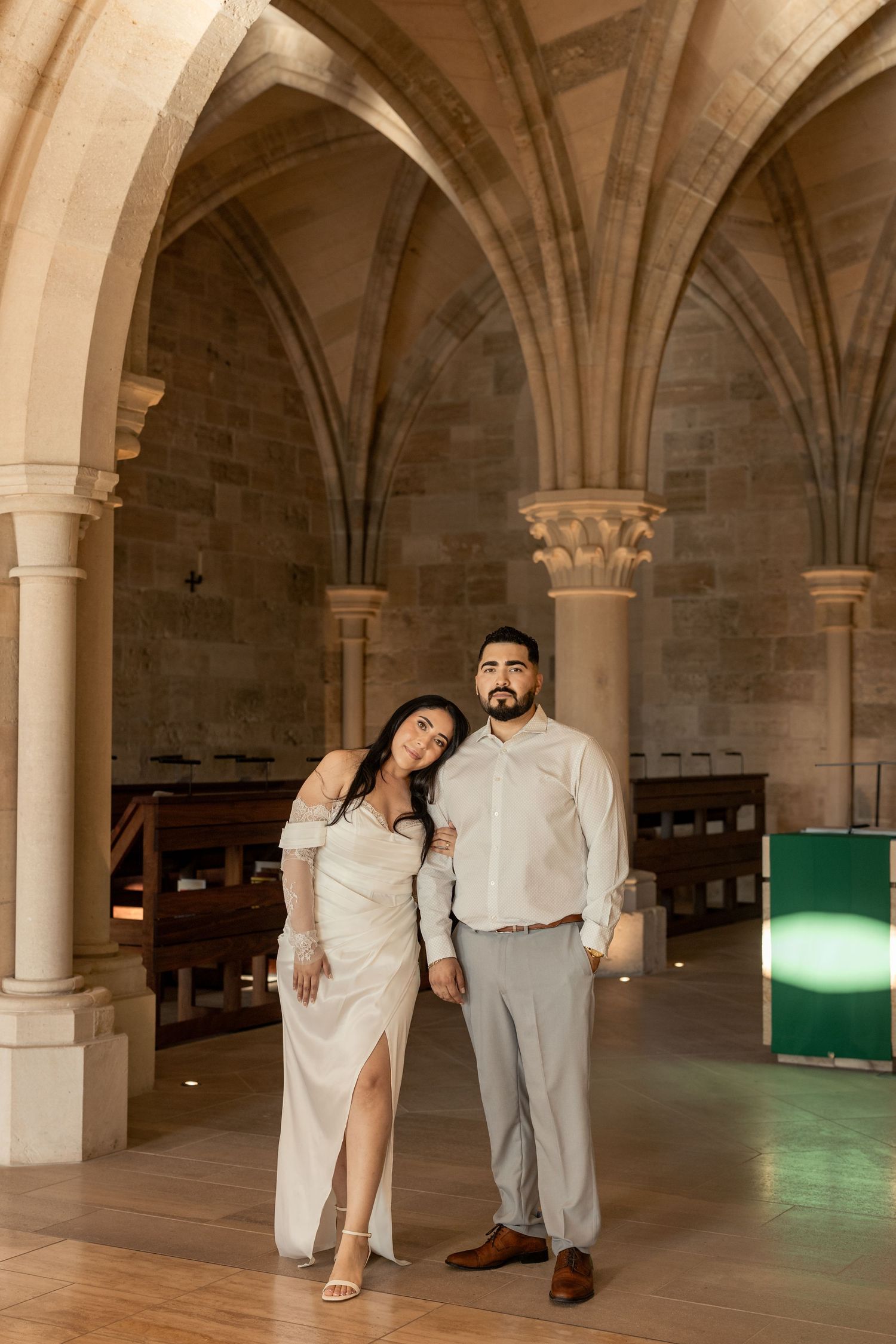 A couple poses together in formal attire inside a historic building with beautiful stone archways.