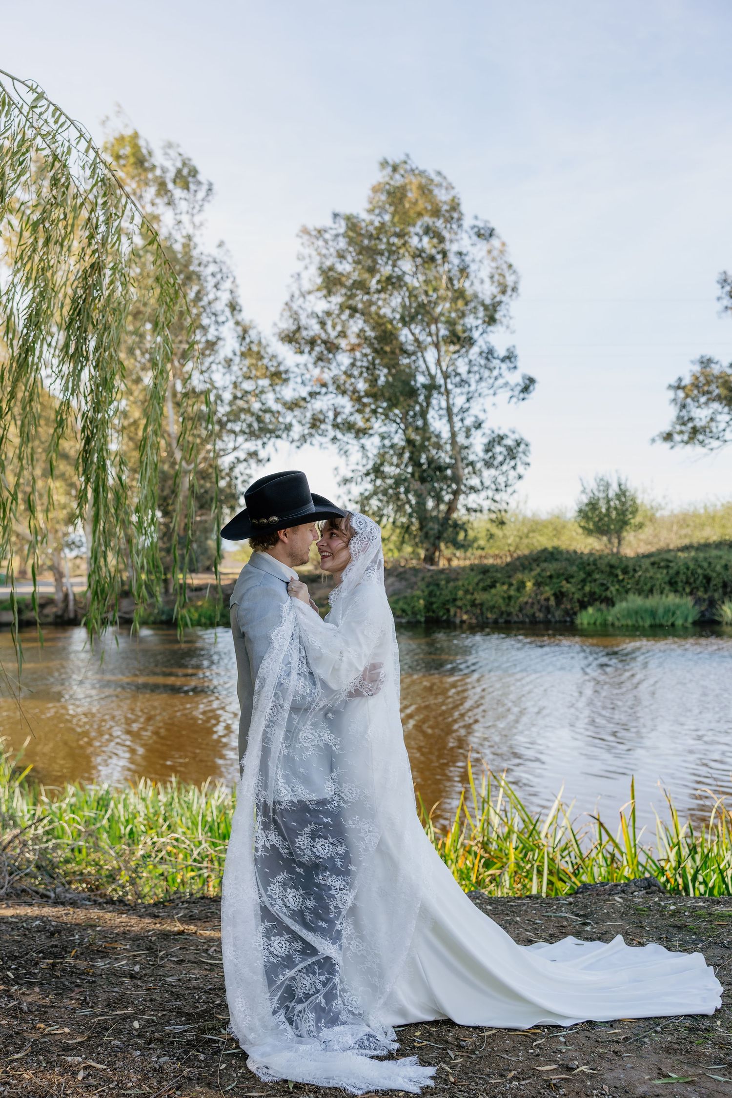 A romantic wedding photo by a lake with willow trees and flowing white dress.