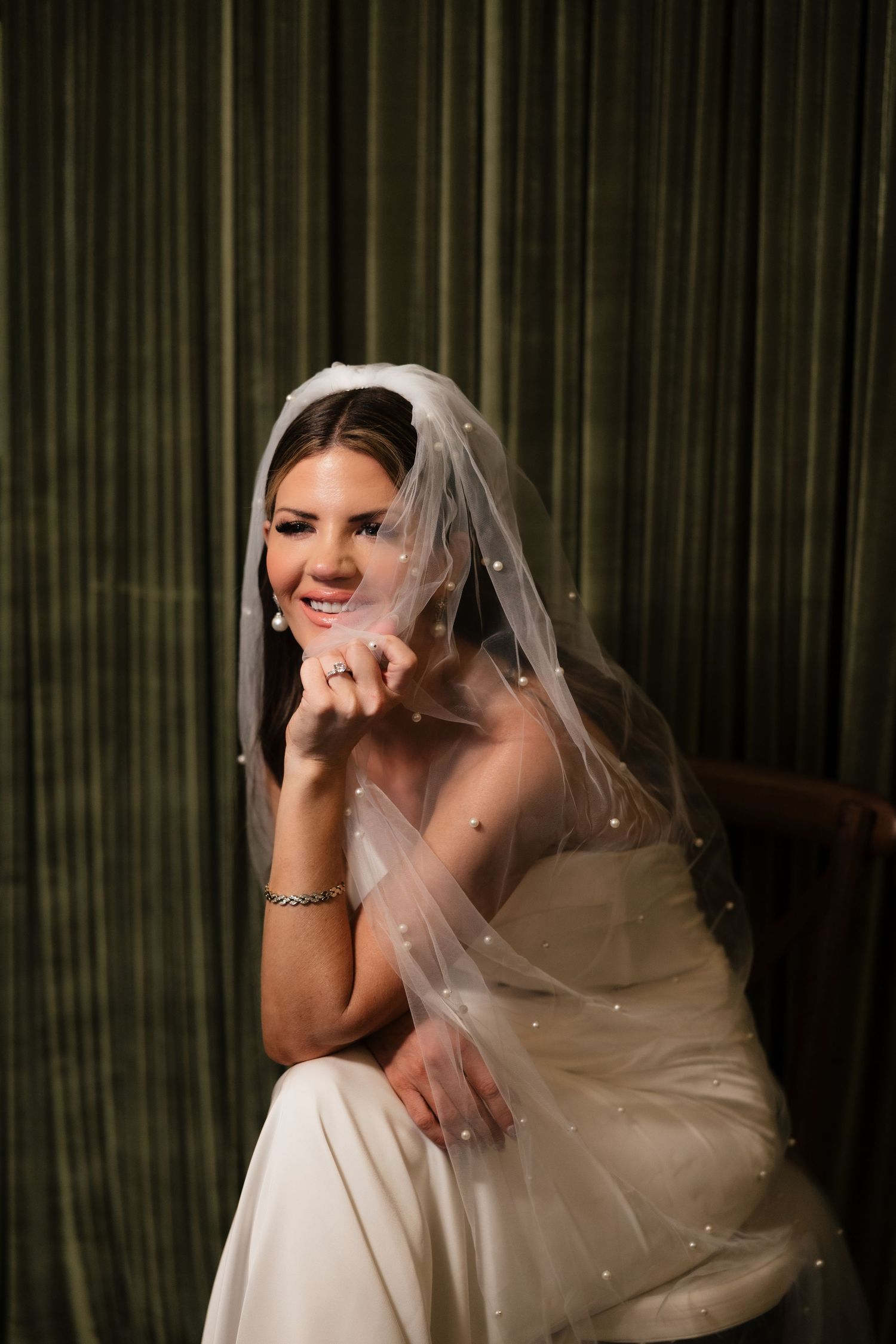 Smiling bride in white gown and pearl-adorned veil sits gracefully, resting chin on hand before green curtains.