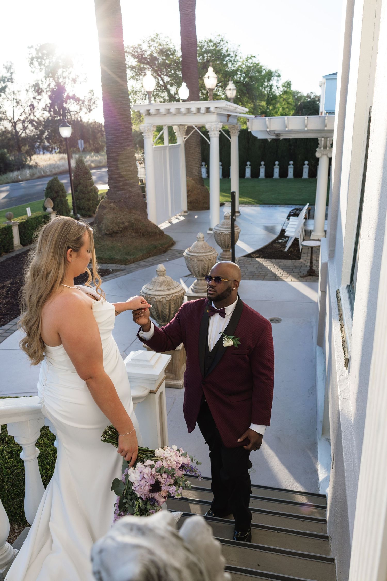 Groom in burgundy tuxedo kisses bride's hand on elegant white porch, bride holding pink bouquet in white gown.