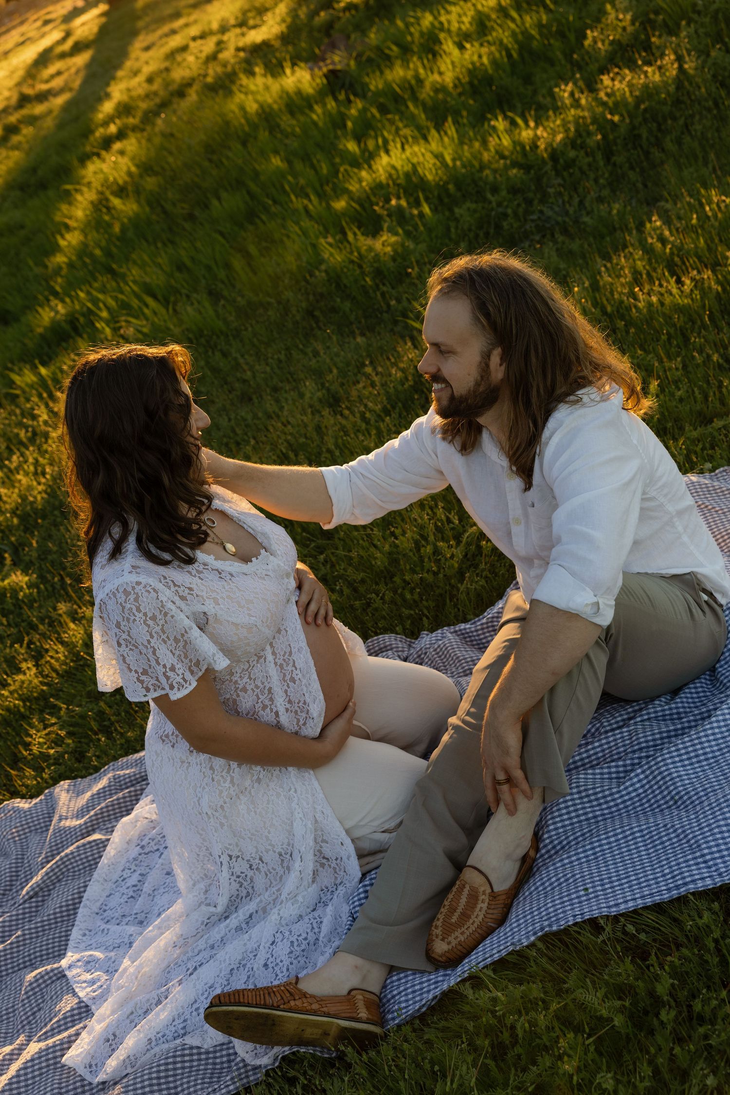 Pregnant couple sitting on blanket in golden sunlight on green grass, man tenderly touching woman in white lace dress.