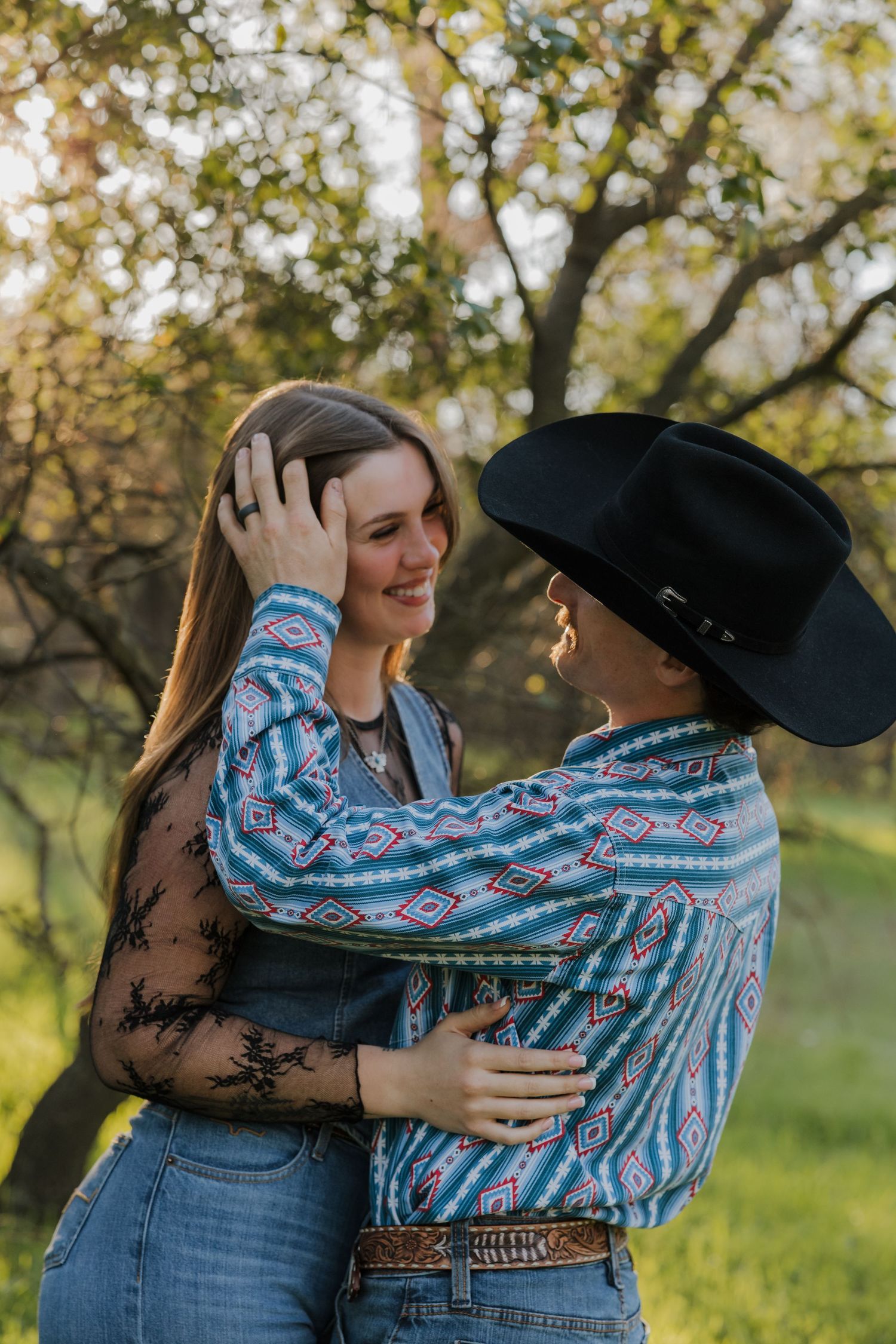 Two women in Western shirts embrace outdoors, one wearing a black cowboy hat, smiling at each other in golden sunlight.