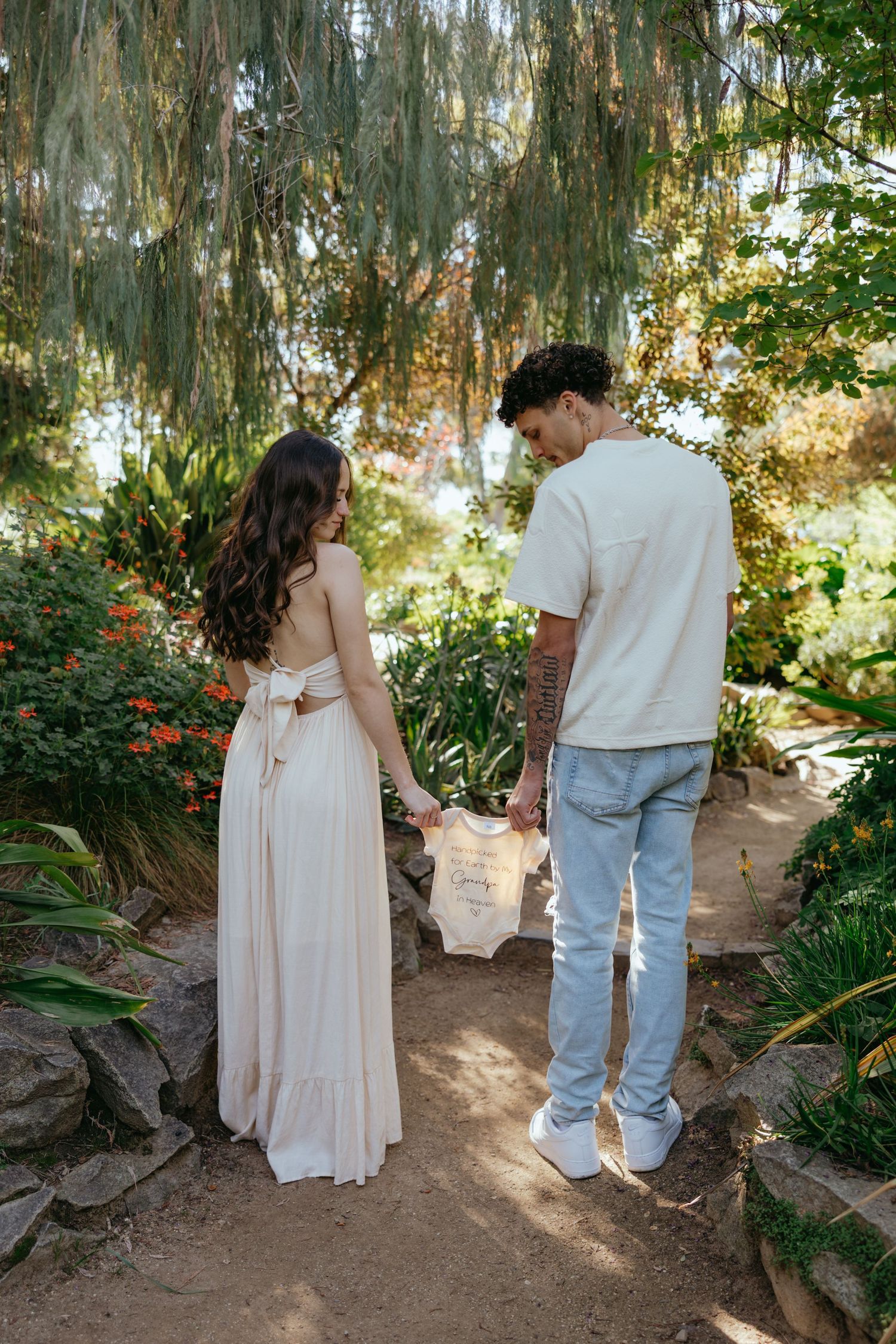Couple holding baby onesie hands on garden path, pregnant maternity photo shoot outdoors.