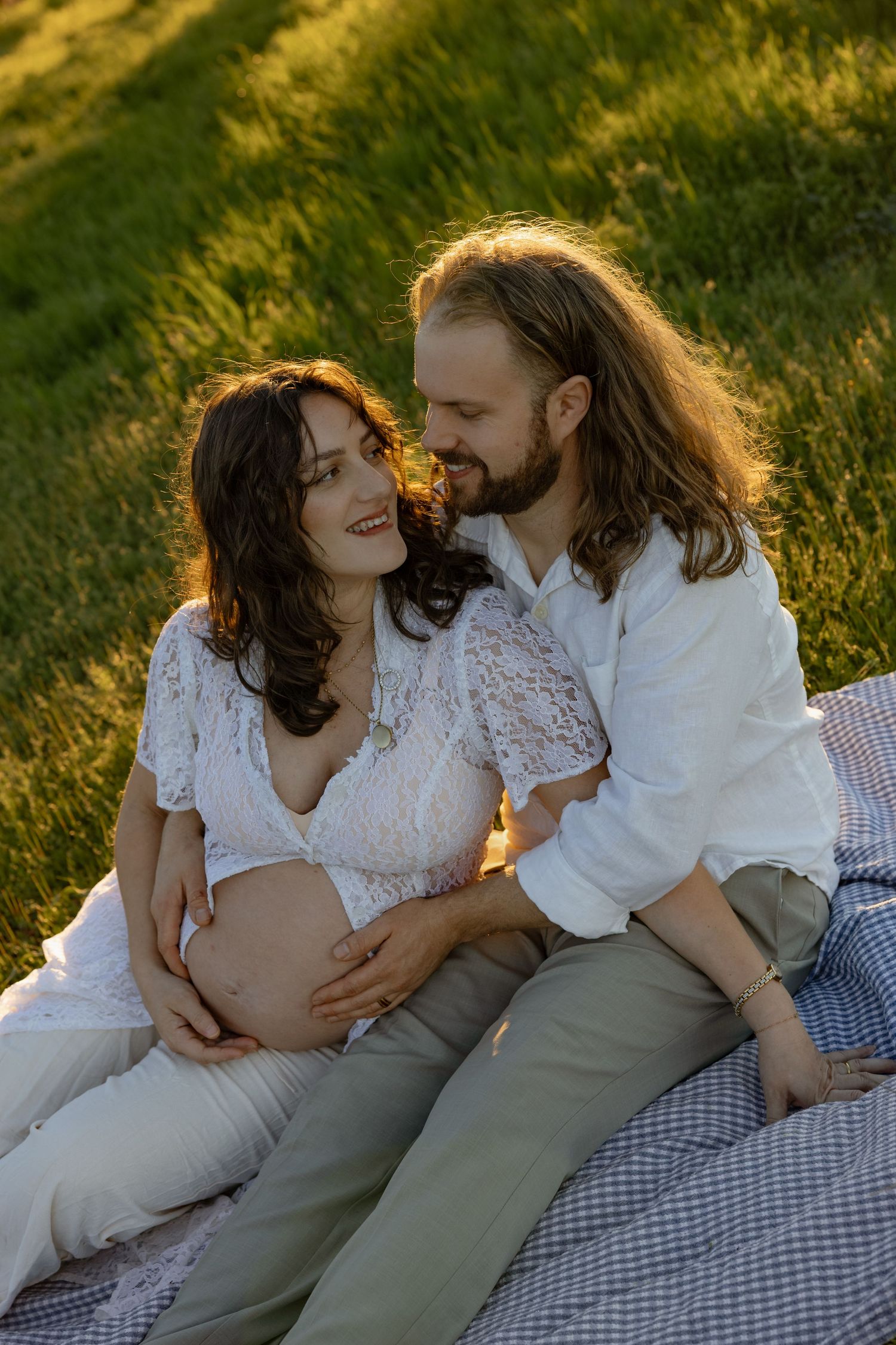 Pregnant couple sitting on a blanket in a green field, sharing a tender moment in golden sunlight.