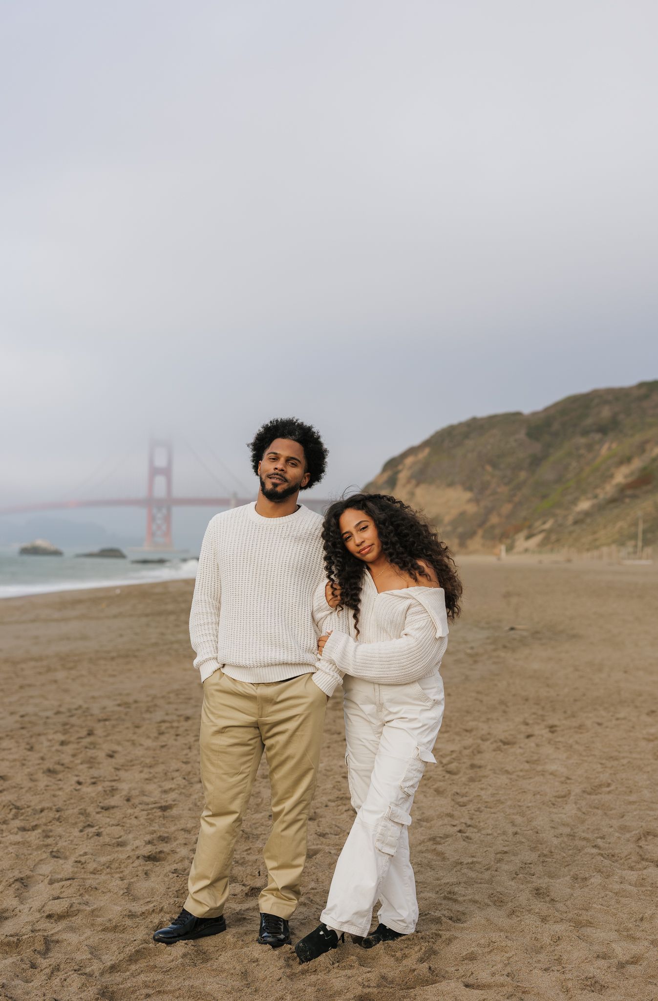A couple in white outfits walks together on a beach with Golden Gate Bridge in background.