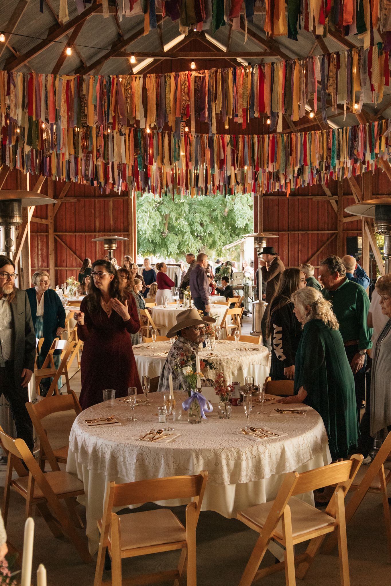 Wedding reception setup inside rustic barn with colorful fabric streamers hanging from ceiling and round tables.