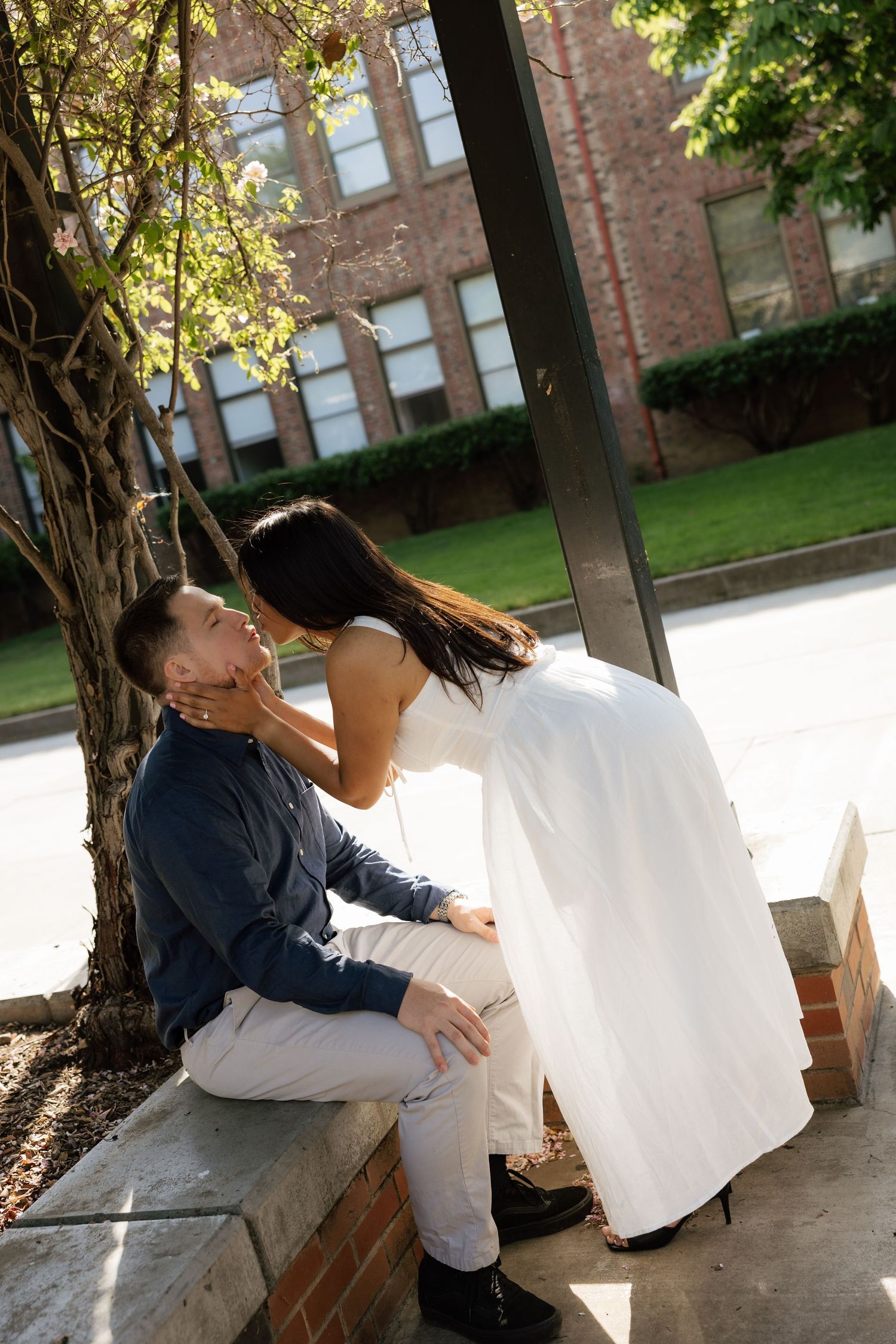 Romantic couple sharing a tender moment outdoors, woman in white dress leaning toward seated man near brick building.
