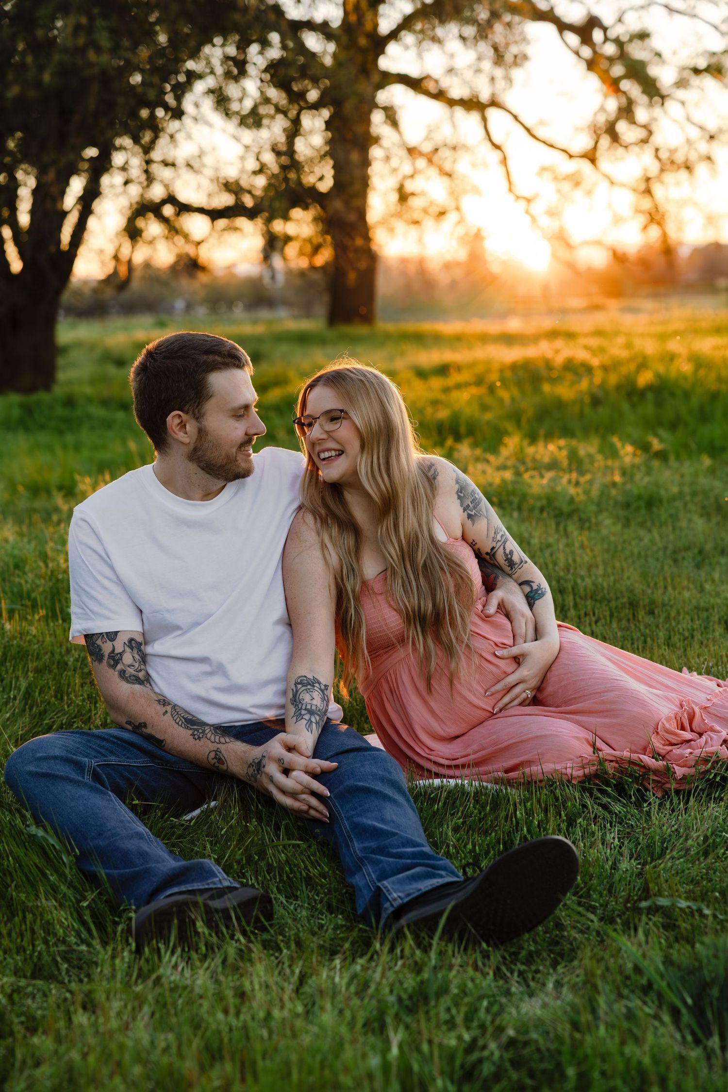 Couple sitting on green grass at golden hour sunset, woman in pink dress and tattooed man in white tee smiling at each other.