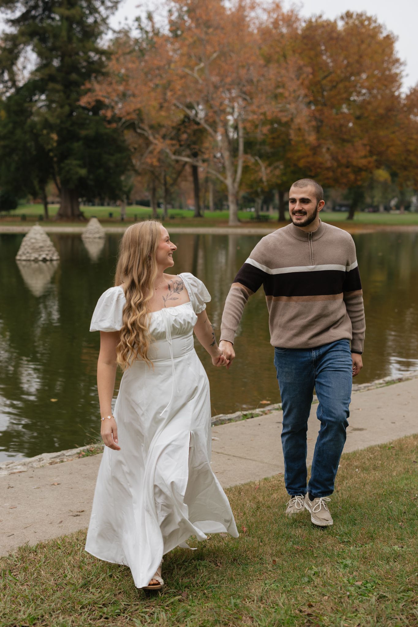Couple walking hand in hand along pond with fall foliage in background.