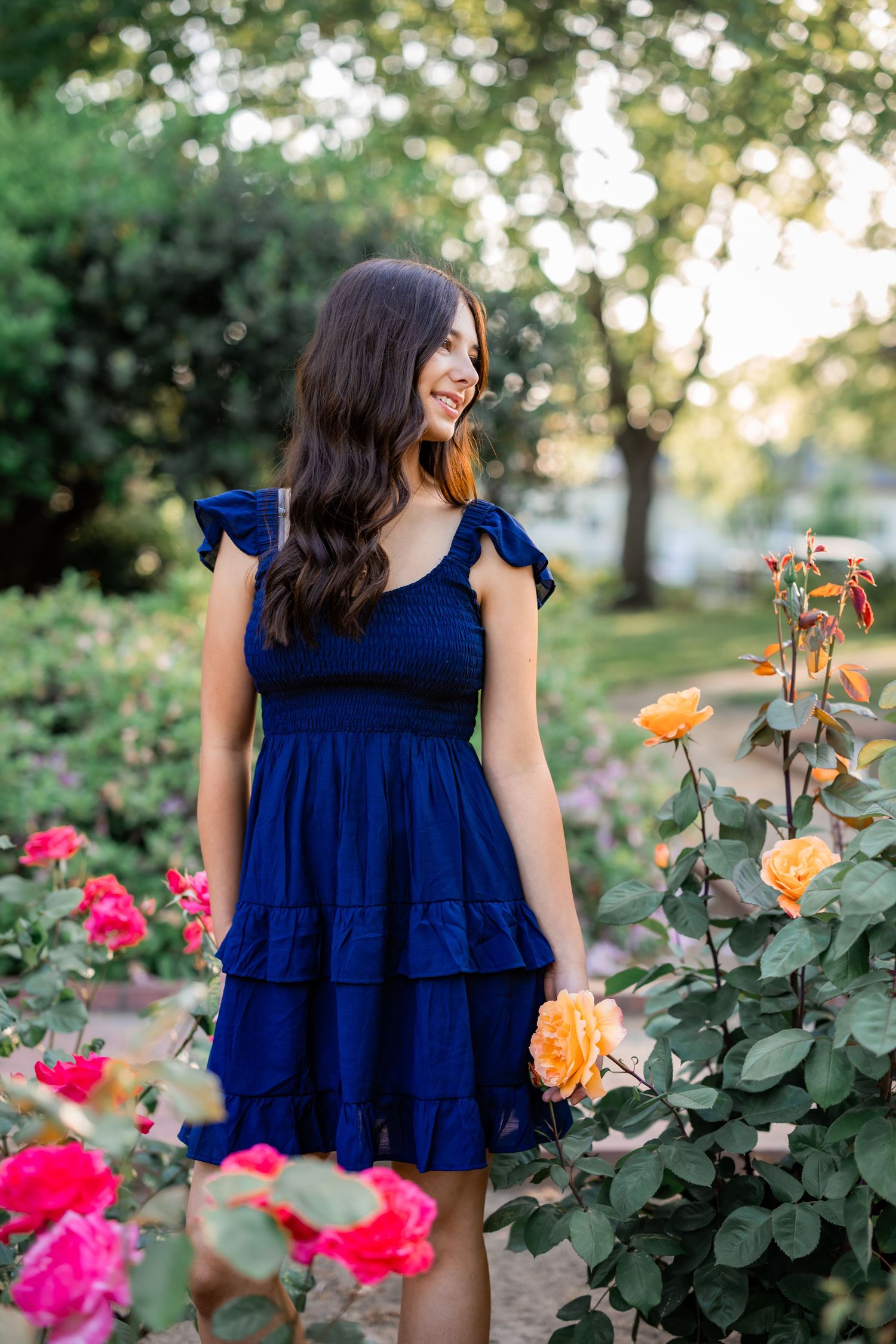 Woman in navy blue ruffle dress poses in garden with blooming roses.