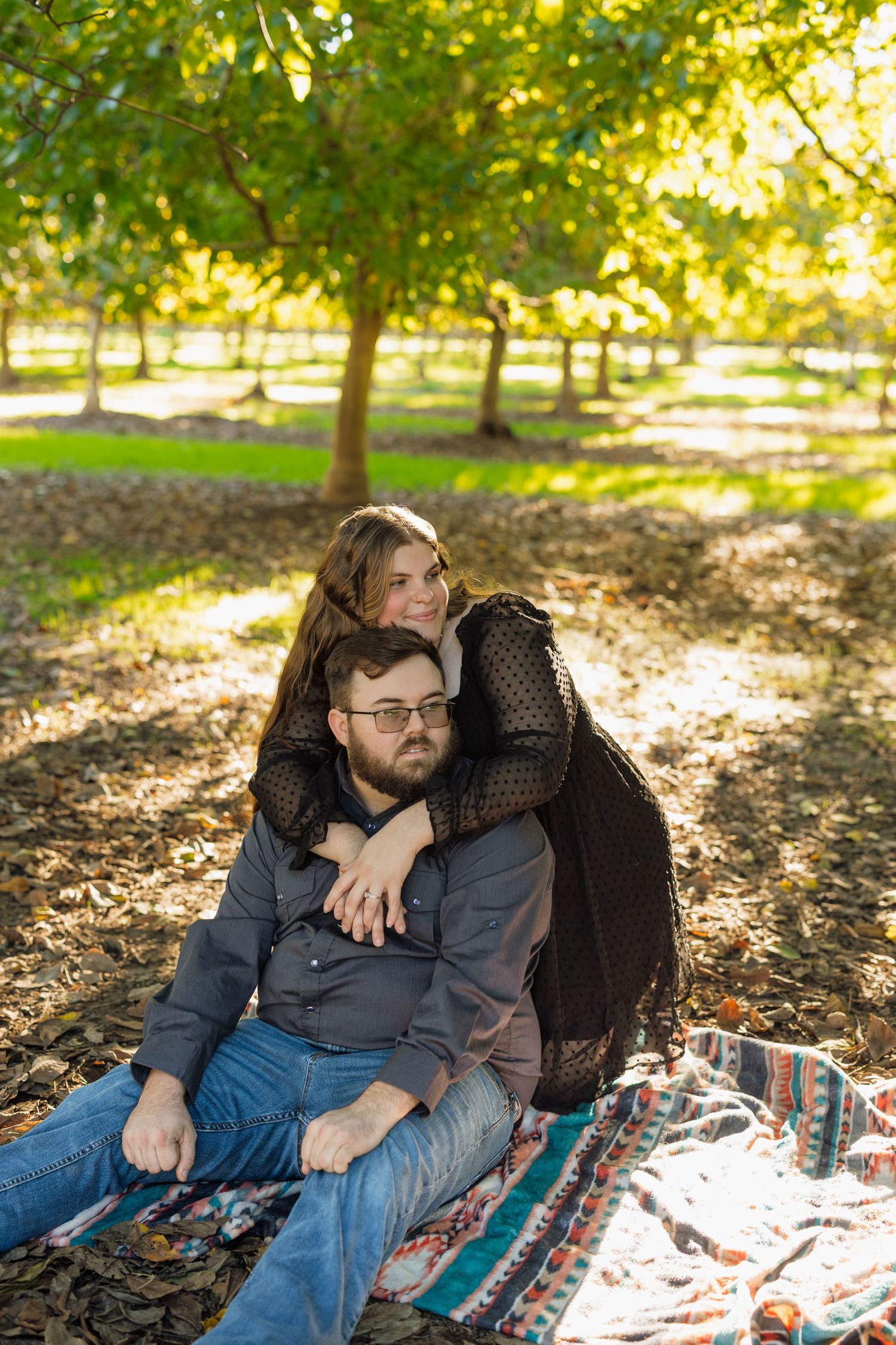 A couple relaxes on a blanket under trees with dappled sunlight filtering through.
