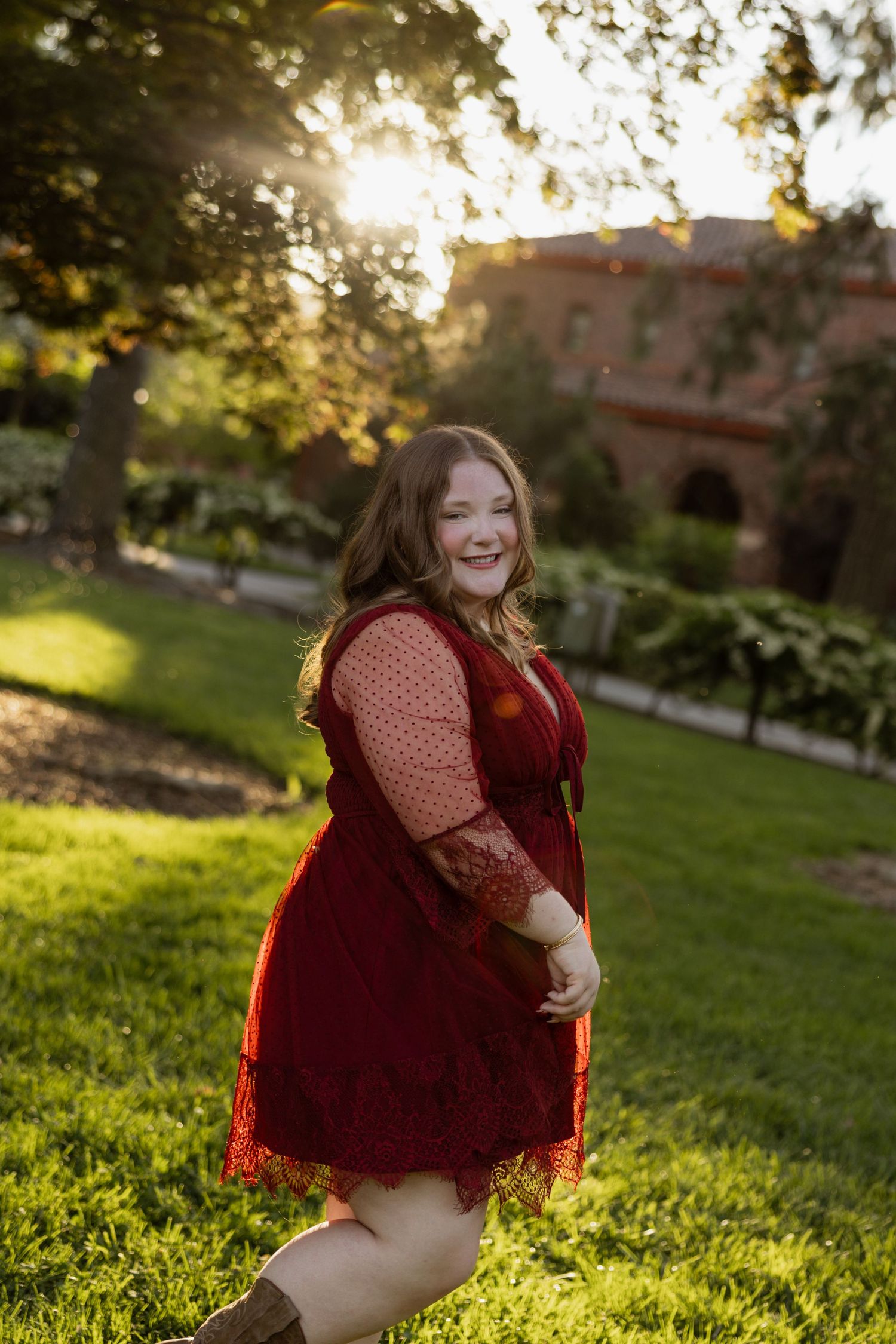 A joyful woman in a red lace dress twirls in a sunlit garden, smiling brightly with golden hour light behind her.