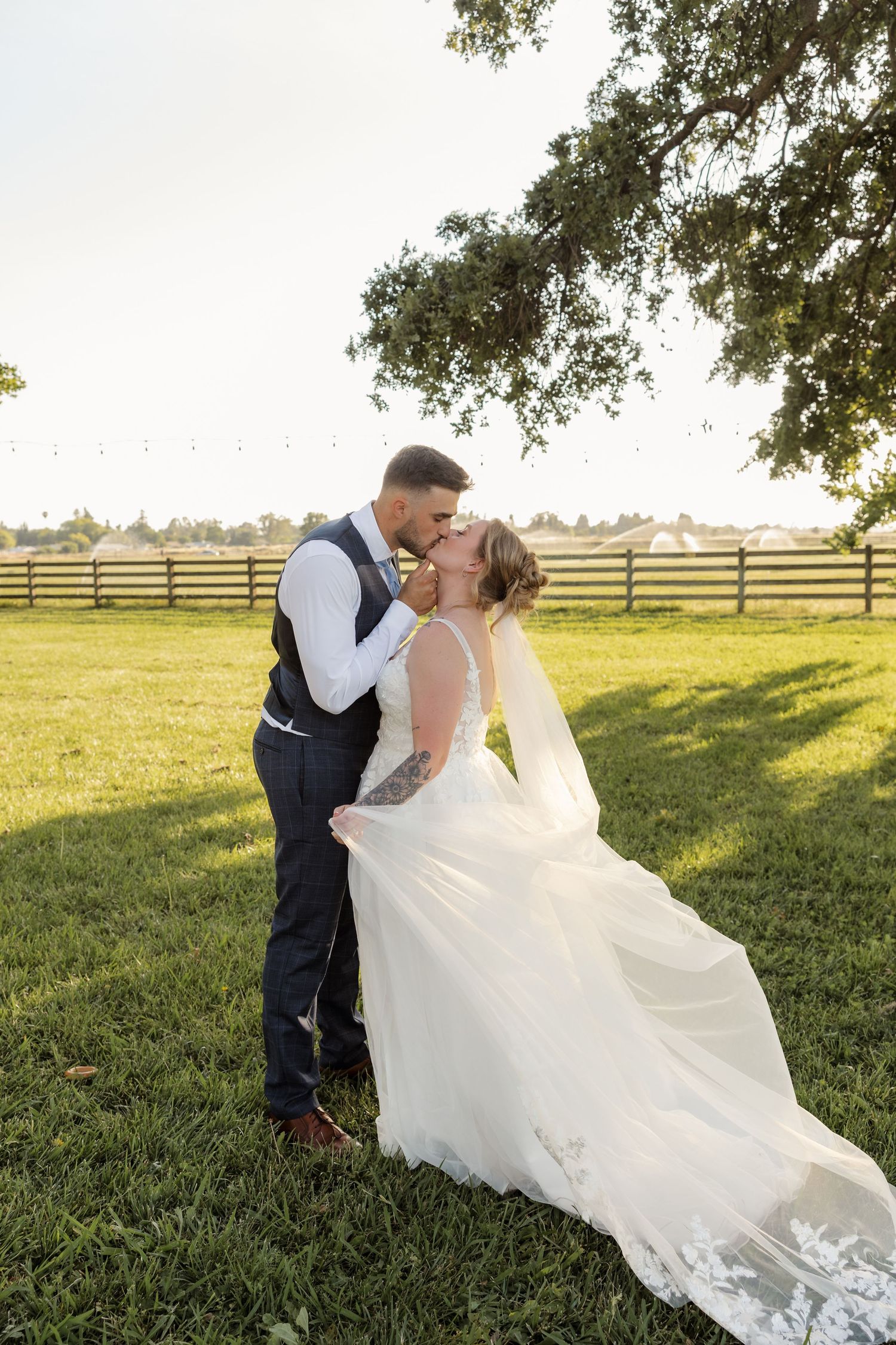 Bride and groom share a romantic kiss on lush green grass at a rustic farm wedding venue during golden hour.