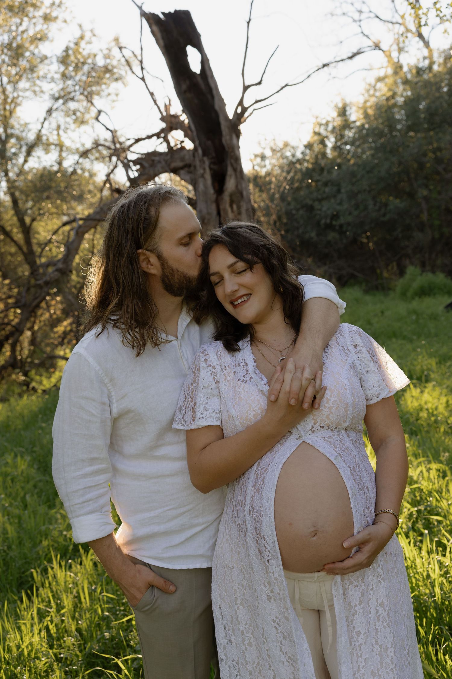 Couple in white outdoors, man kissing pregnant woman's cheek while she cradles her baby bump in a sunny green field.