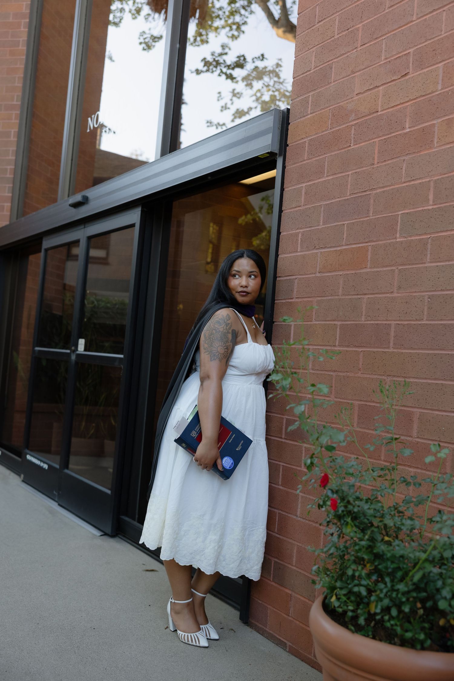 A person in a white dress stands near a brick building with windows and small garden plants.