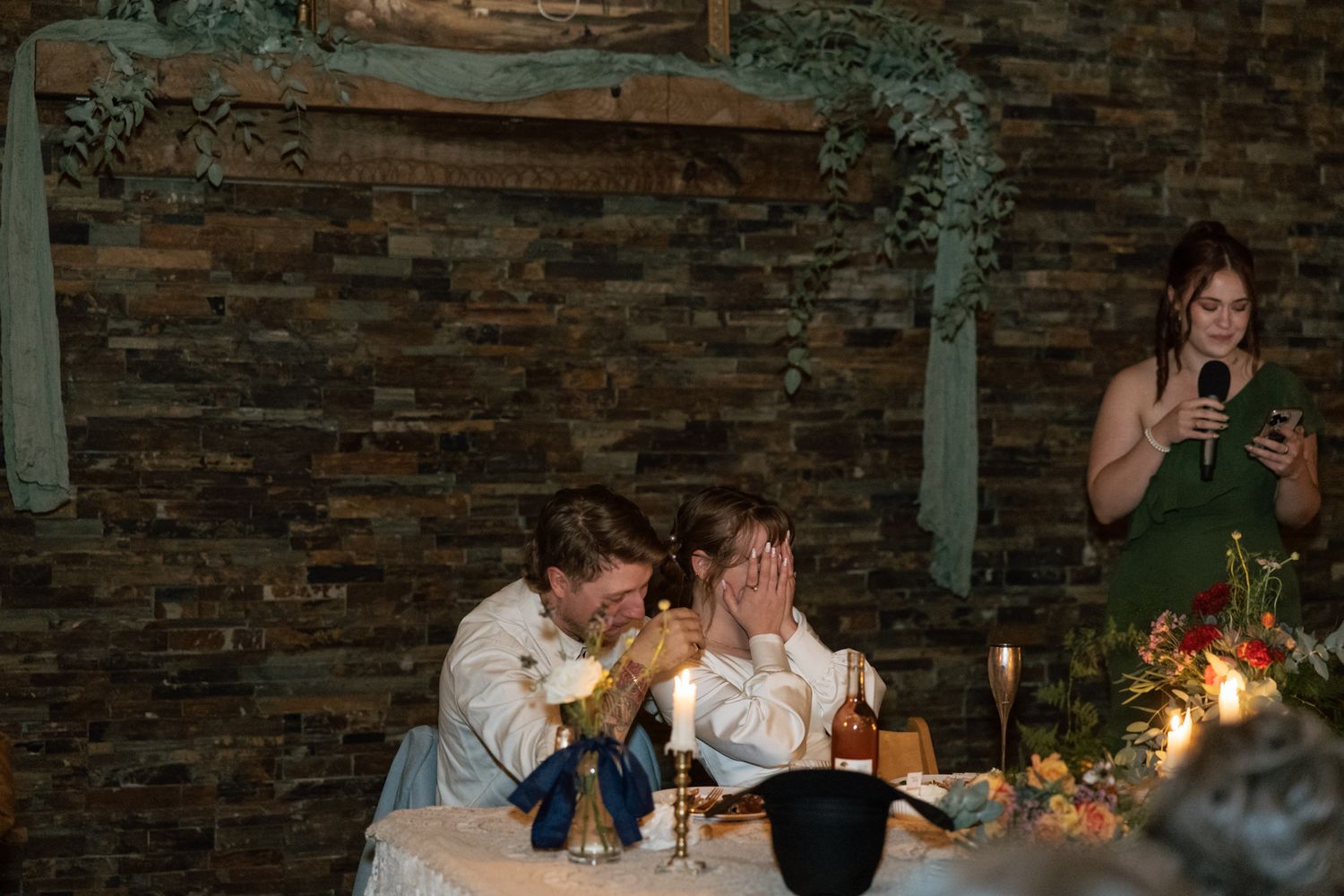 Candlelit reception table at rustic venue with stone wall backdrop and floral centerpieces.