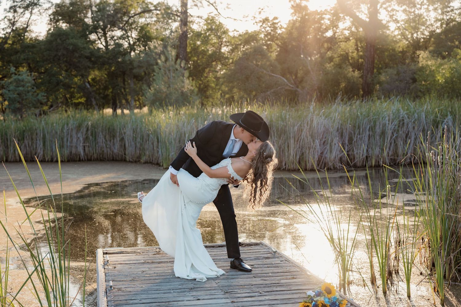 Couple sharing a romantic dip kiss on a wooden dock by a sunlit pond, bride in white gown and groom in black hat.