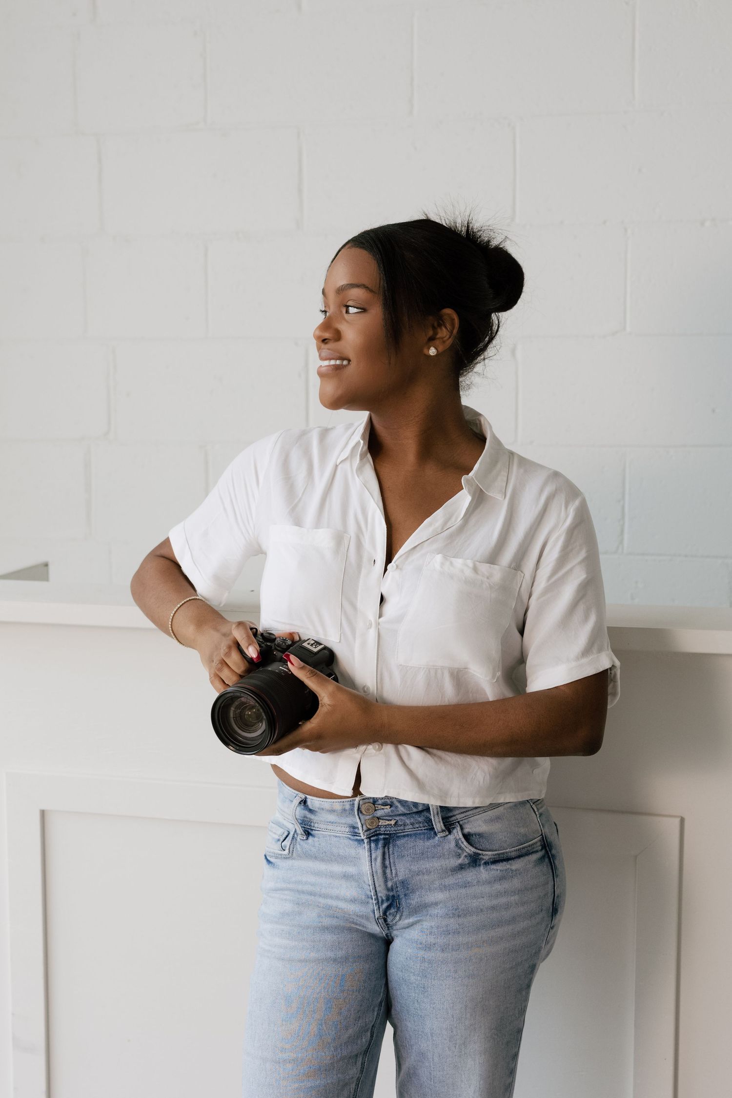 Professional photographer in white button-down shirt and jeans poses against white wall with camera in studio.