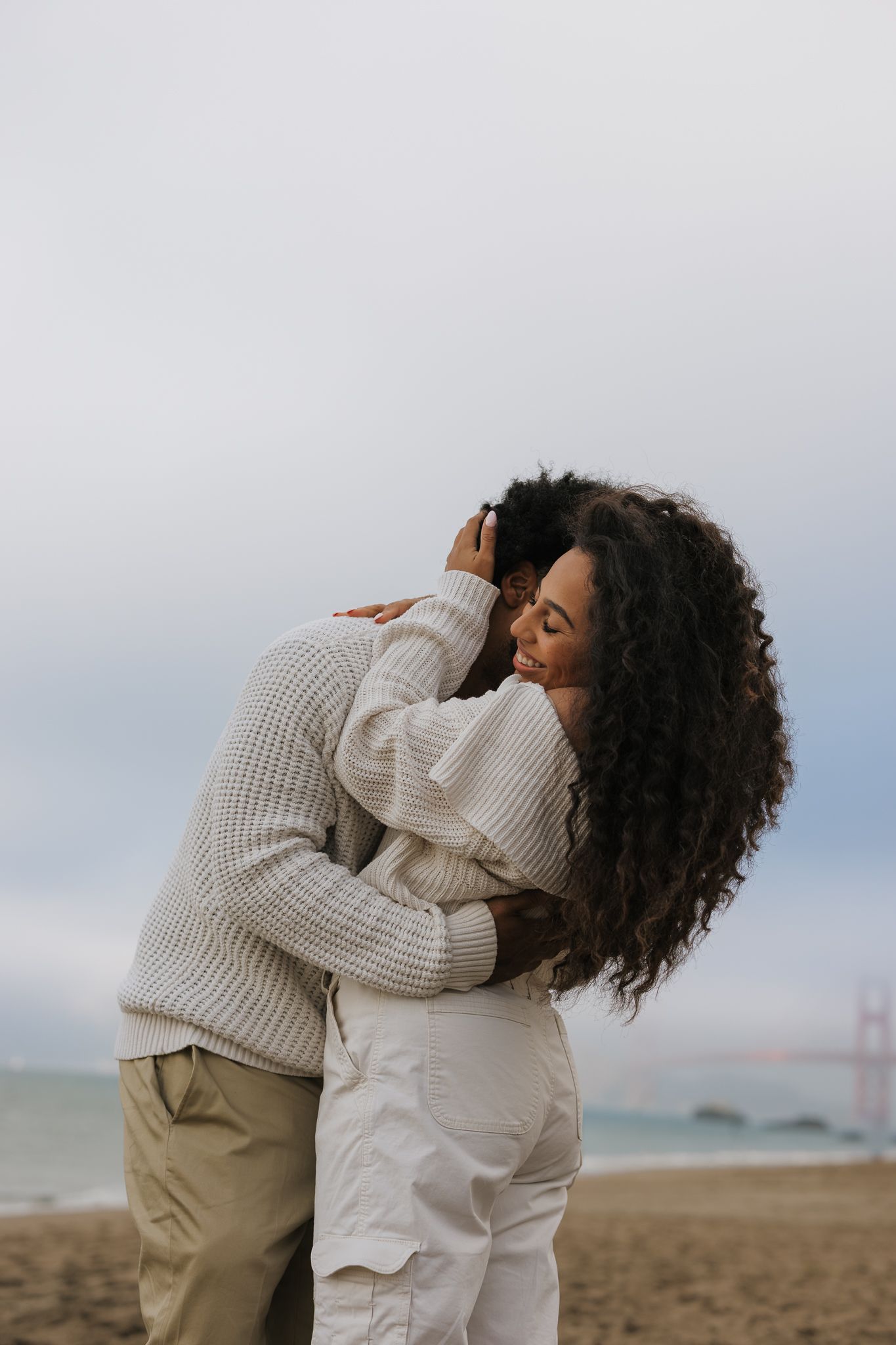 Intimate embrace on beach with sweaters and cloudy coastal backdrop.