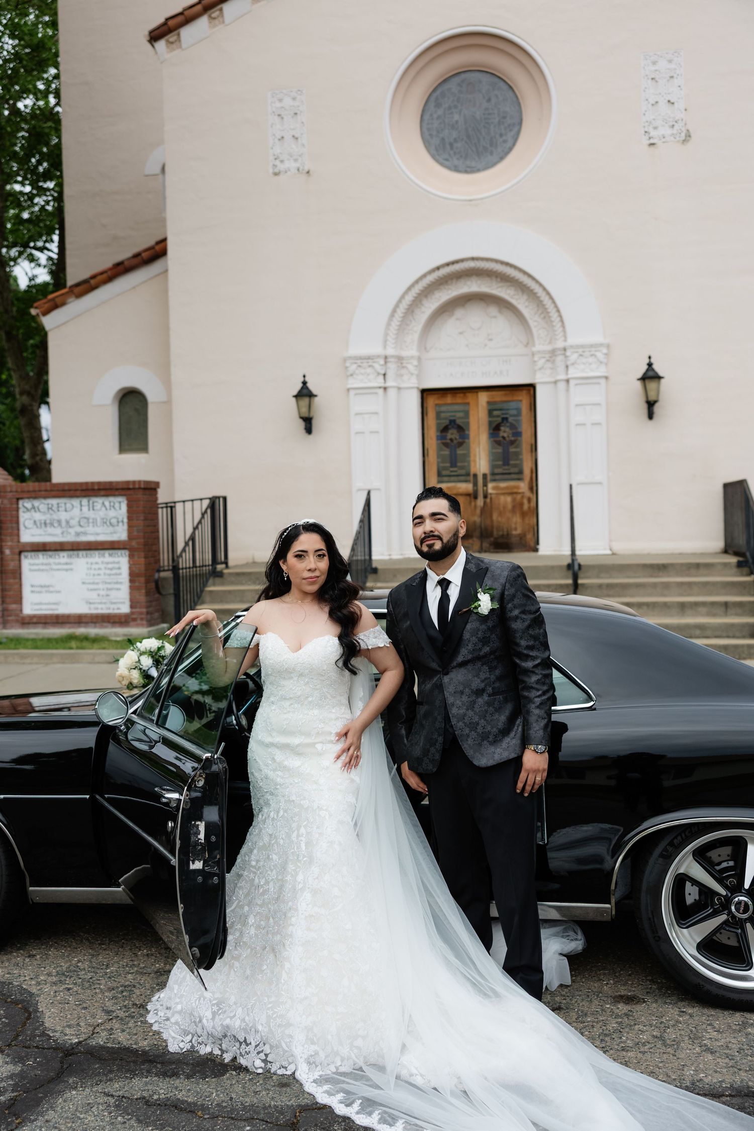 Bride in white mermaid gown and groom in dark suit pose by classic black car outside Sacred Heart Catholic Church.