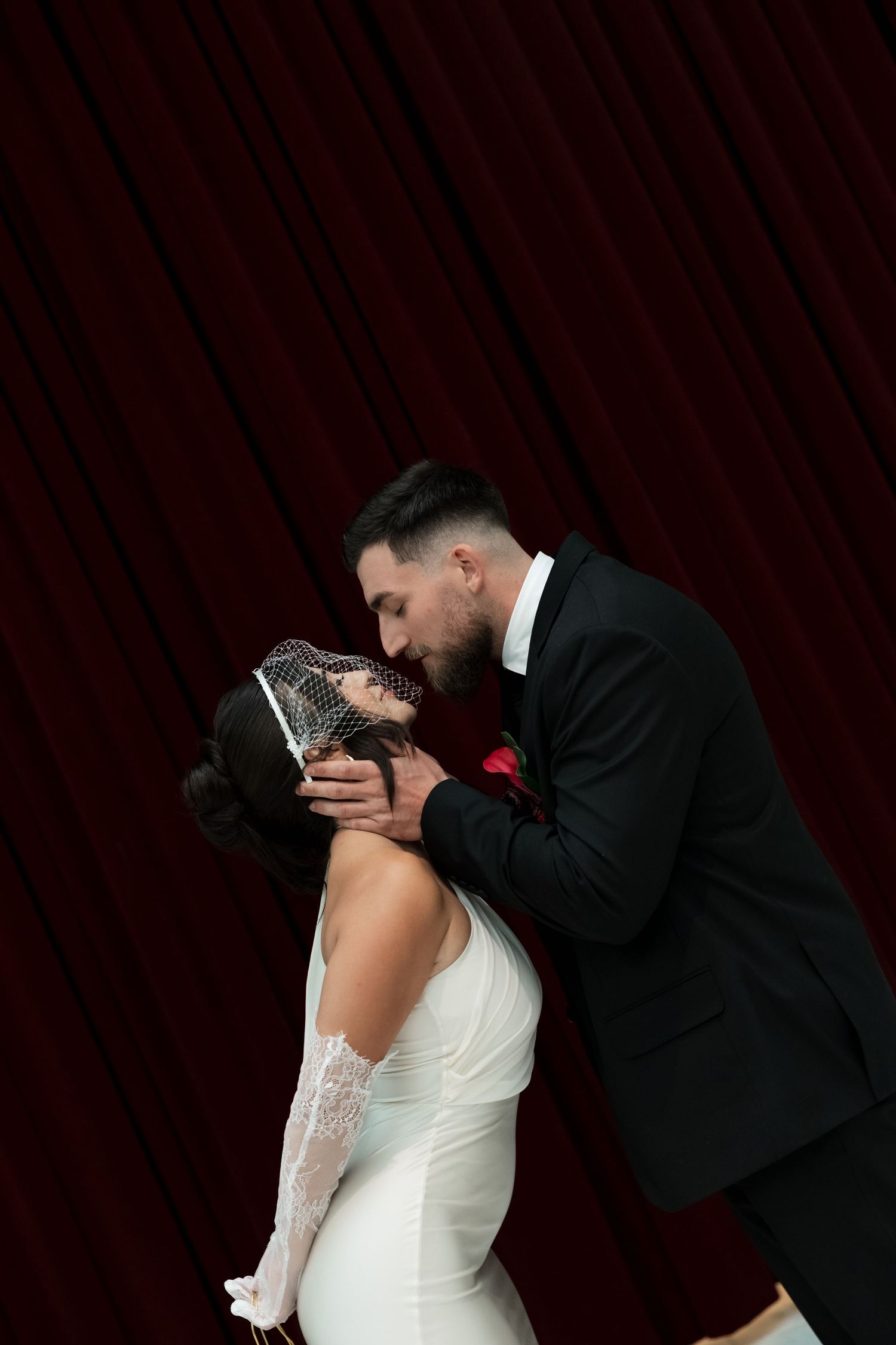 A bride and groom share a romantic kiss in front of a deep red curtain backdrop on their wedding day.