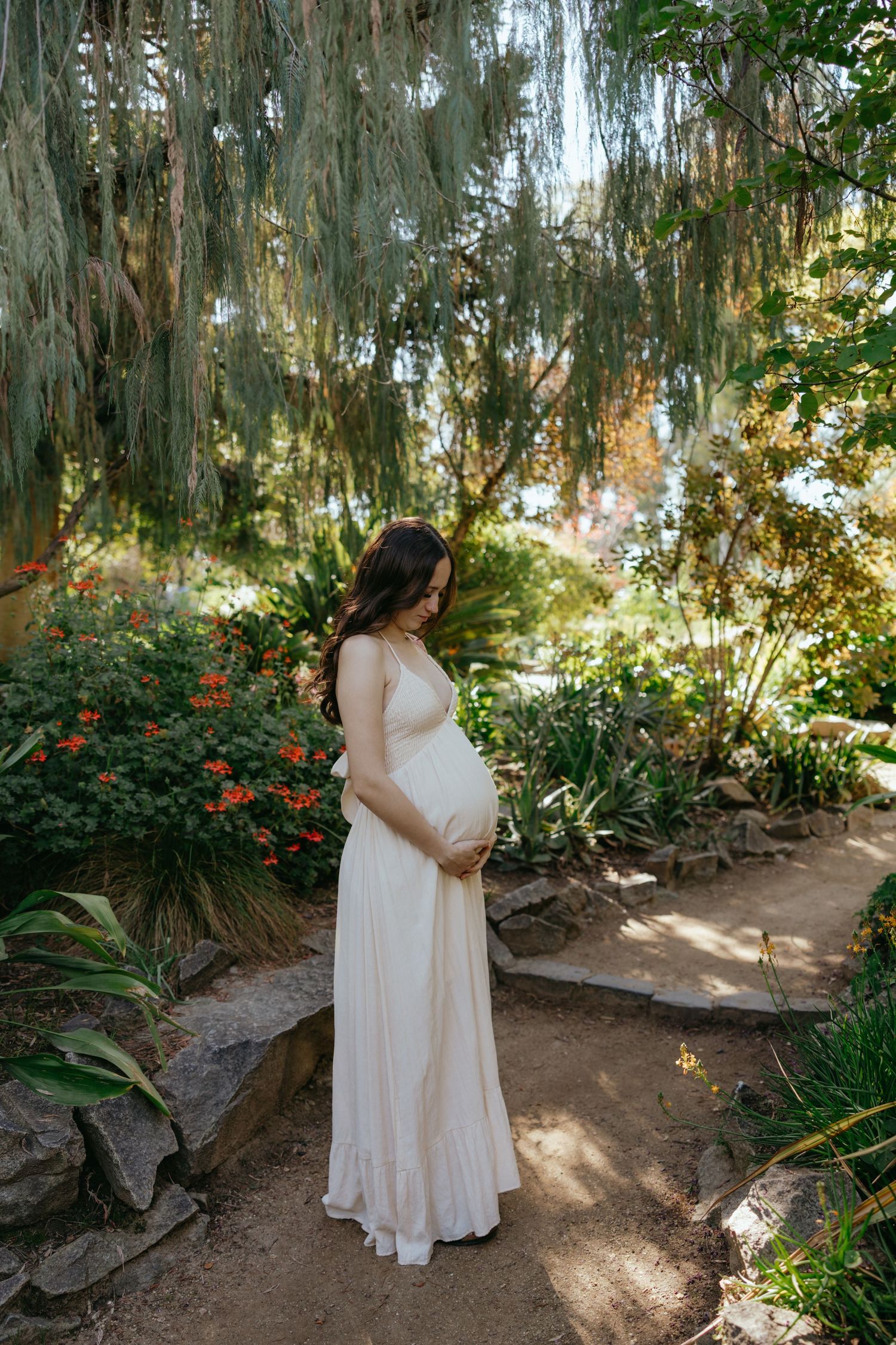 Pregnant woman in white dress cradling baby bump in lush garden, maternity photography.