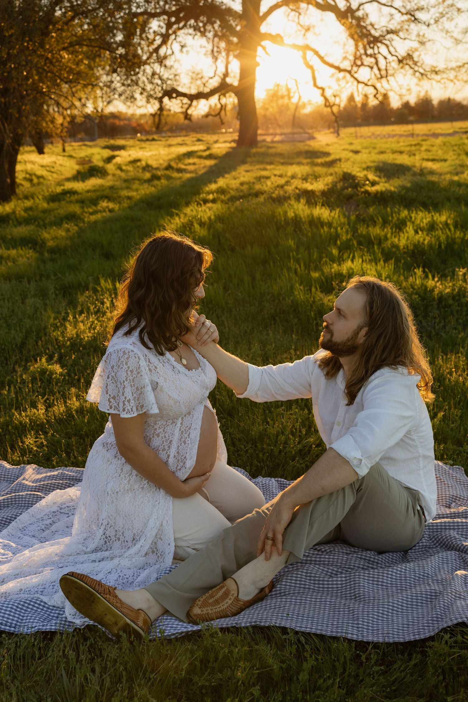 Pregnant couple sitting on blanket in golden sunset light, man tenderly touching woman's face in green meadow.