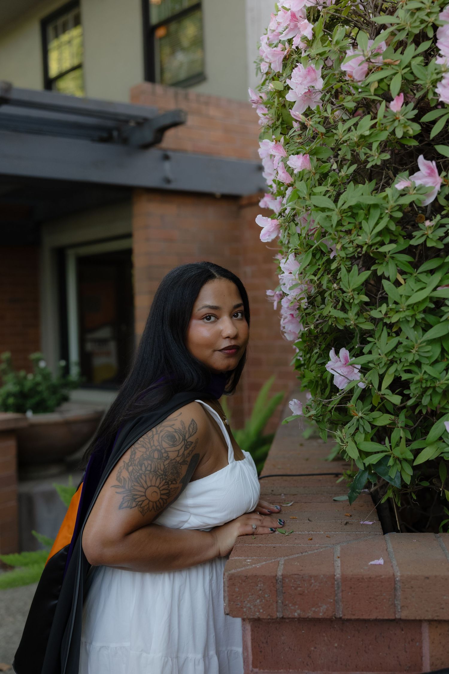 A person in a white dress poses thoughtfully next to pink flowering vines growing on brick steps outdoors.