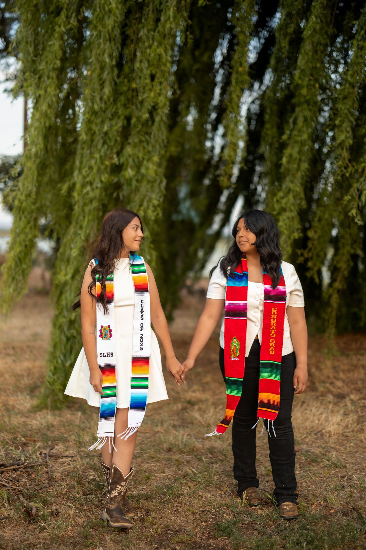 Two people wearing colorful graduation stoles walking together under willow trees.