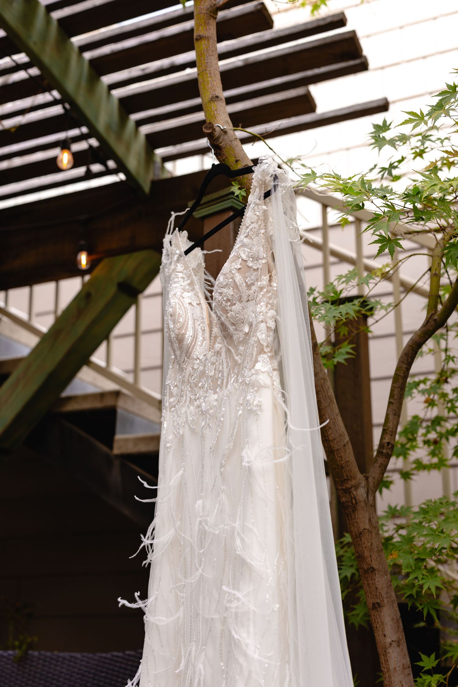 Delicate white lace wedding dress hanging from tree branch under wooden pergola.