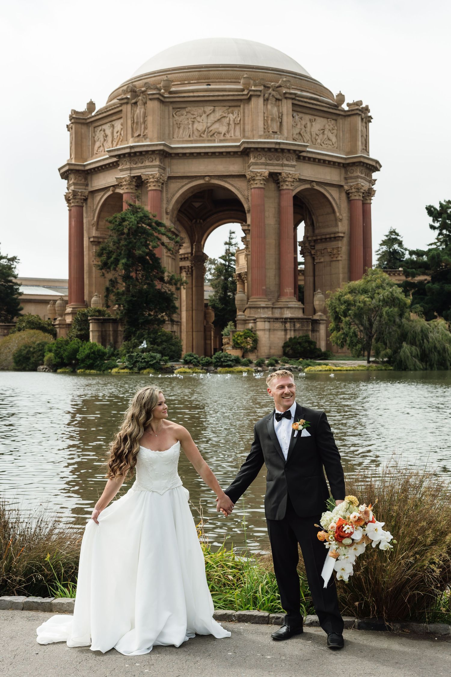Bride and groom hold hands at Palace of Fine Arts in San Francisco, with lagoon and rotunda backdrop.