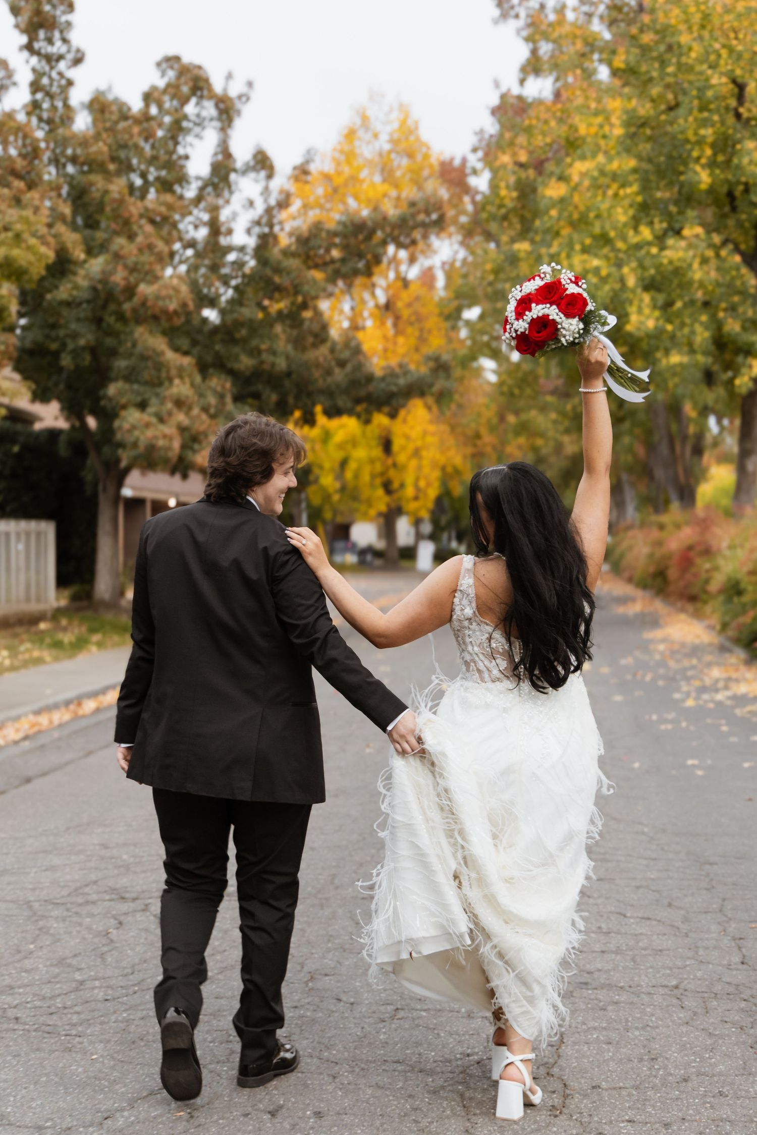 Playful newlyweds walking and dancing together on a tree-lined autumn path.