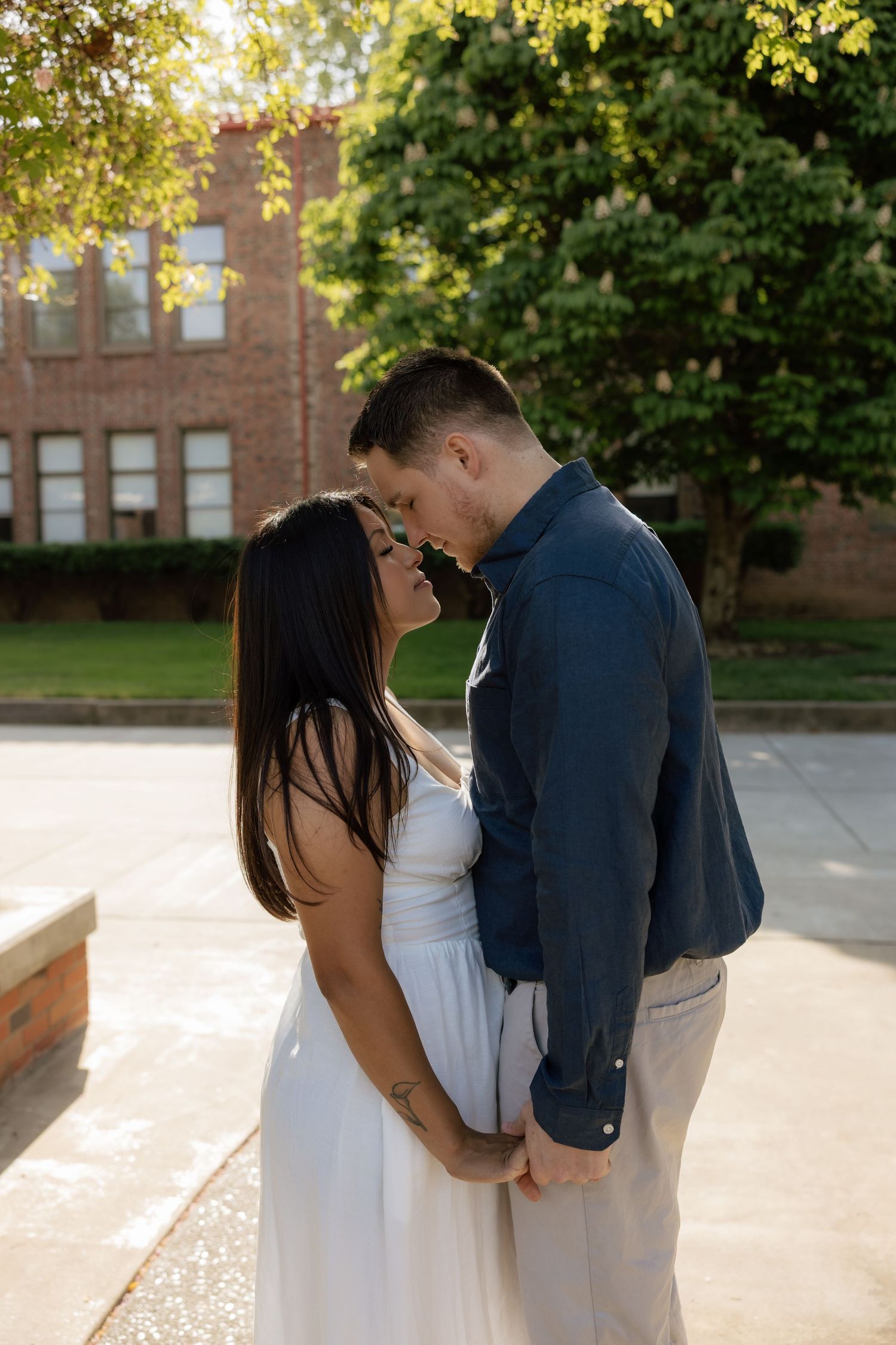Couple sharing a romantic moment outdoors, woman in white dress and man in navy shirt, with brick building backdrop.