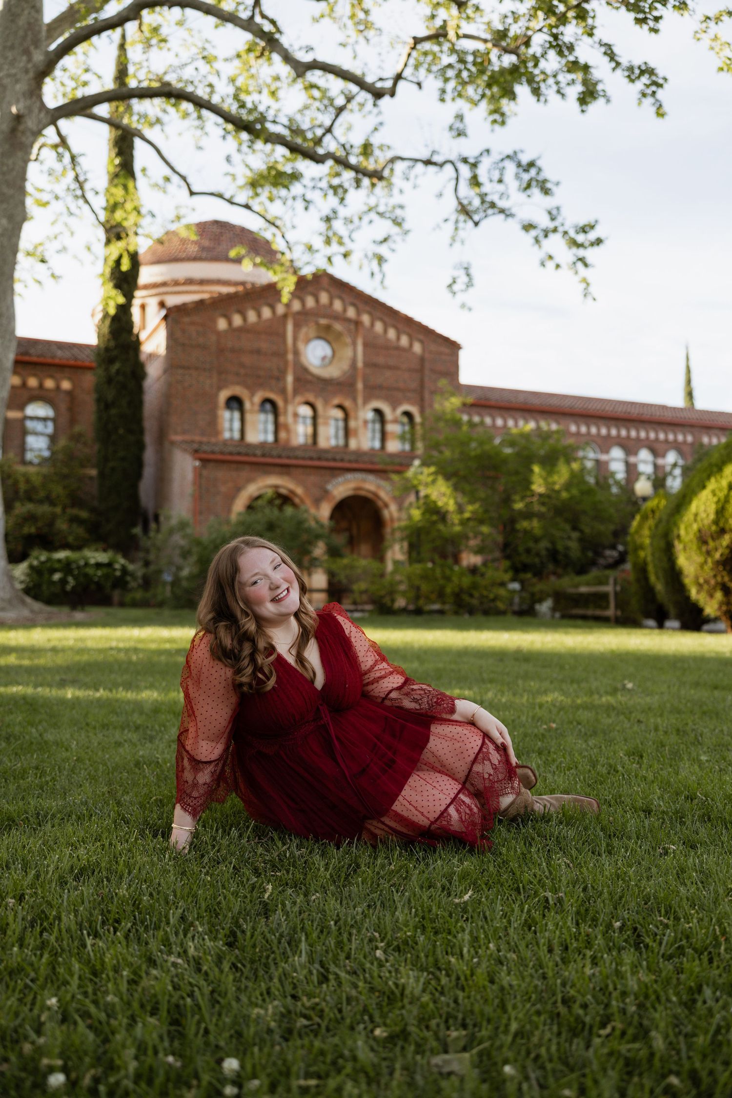 Young woman in red lace dress sitting on green grass, smiling, with historic brick building in background.