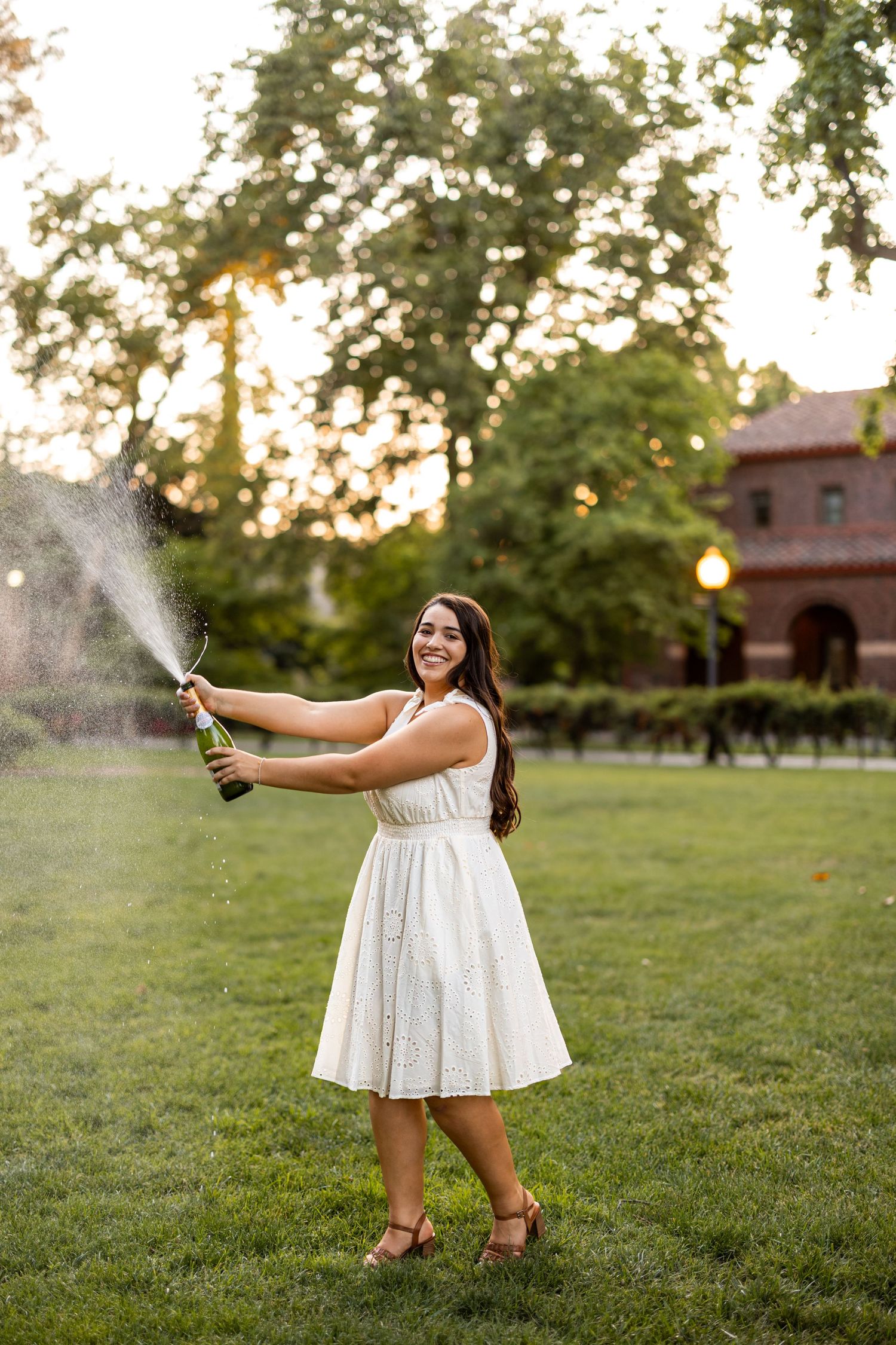 A person in a white dress sprays champagne in a grassy field during golden hour.