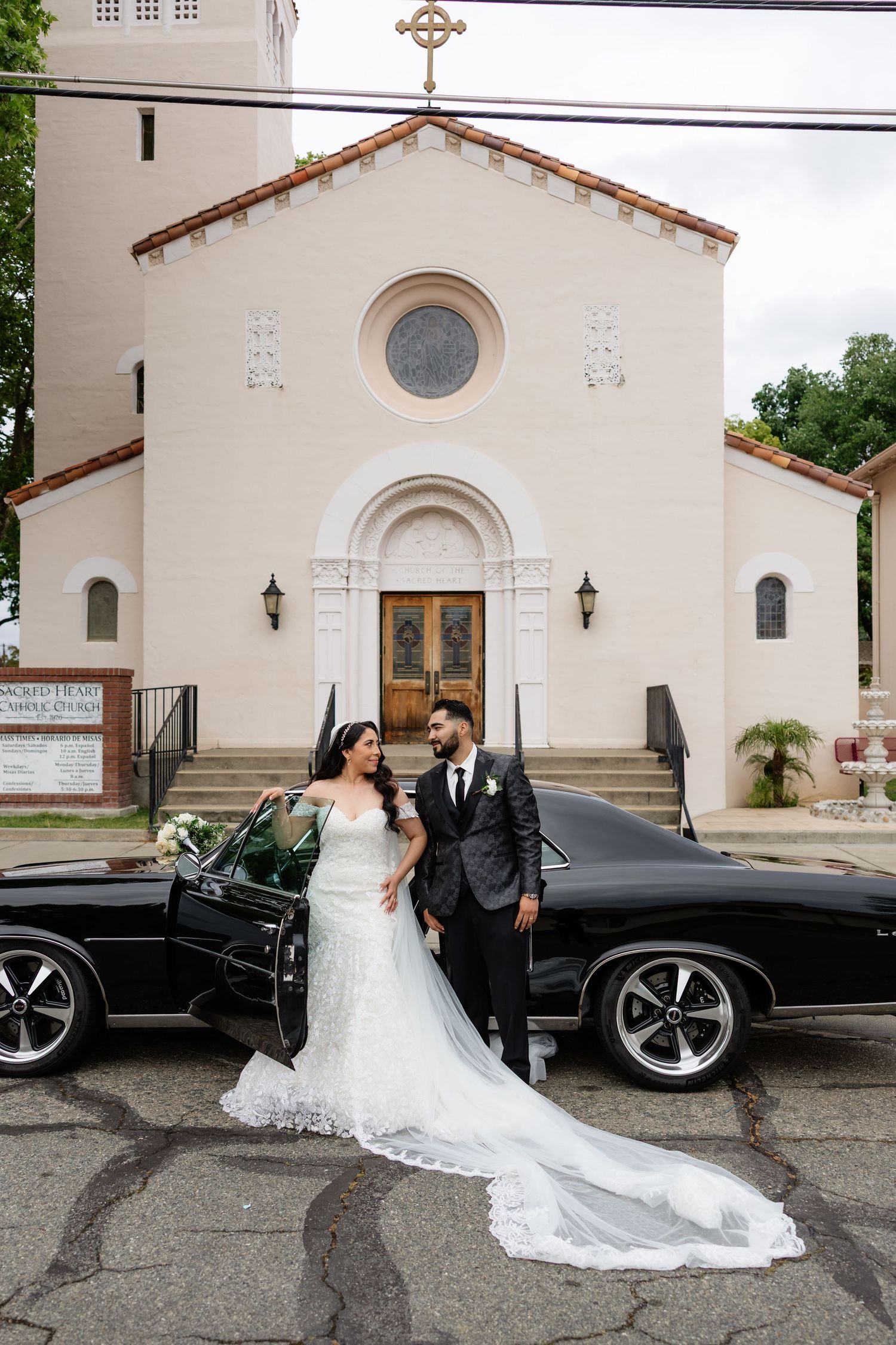 Bride and groom pose beside a classic black car in front of a white church after their wedding ceremony.